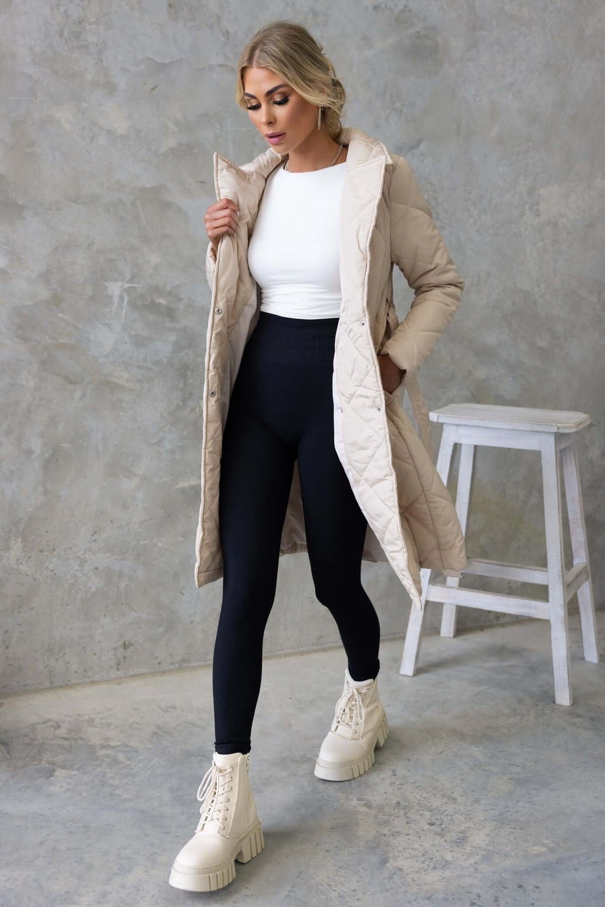 Model wearing a beige longline quilted coat over a white top and black leggings, showcasing a stylish winter outfit.