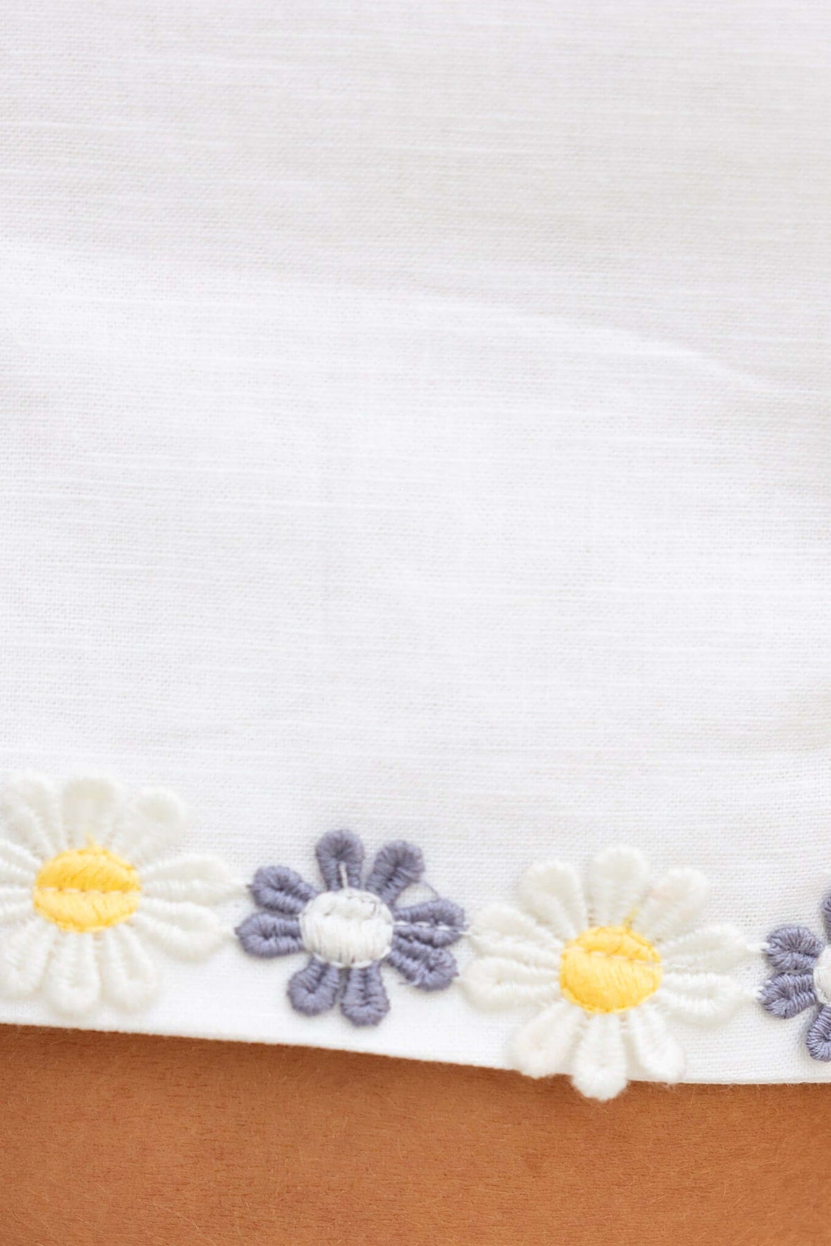 Close-up of floral embroidery on the hem of a white linen top with yellow and purple daisies.