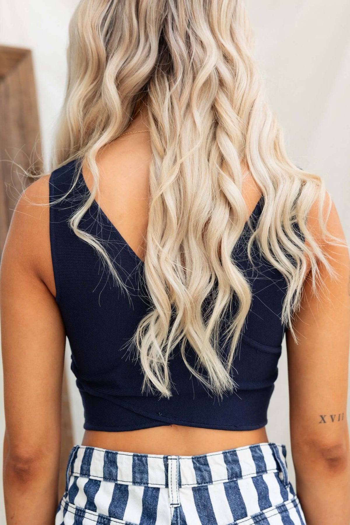 Back view of a woman wearing a navy Orem Top with V-neckline, styled with wavy hair and striped pants.