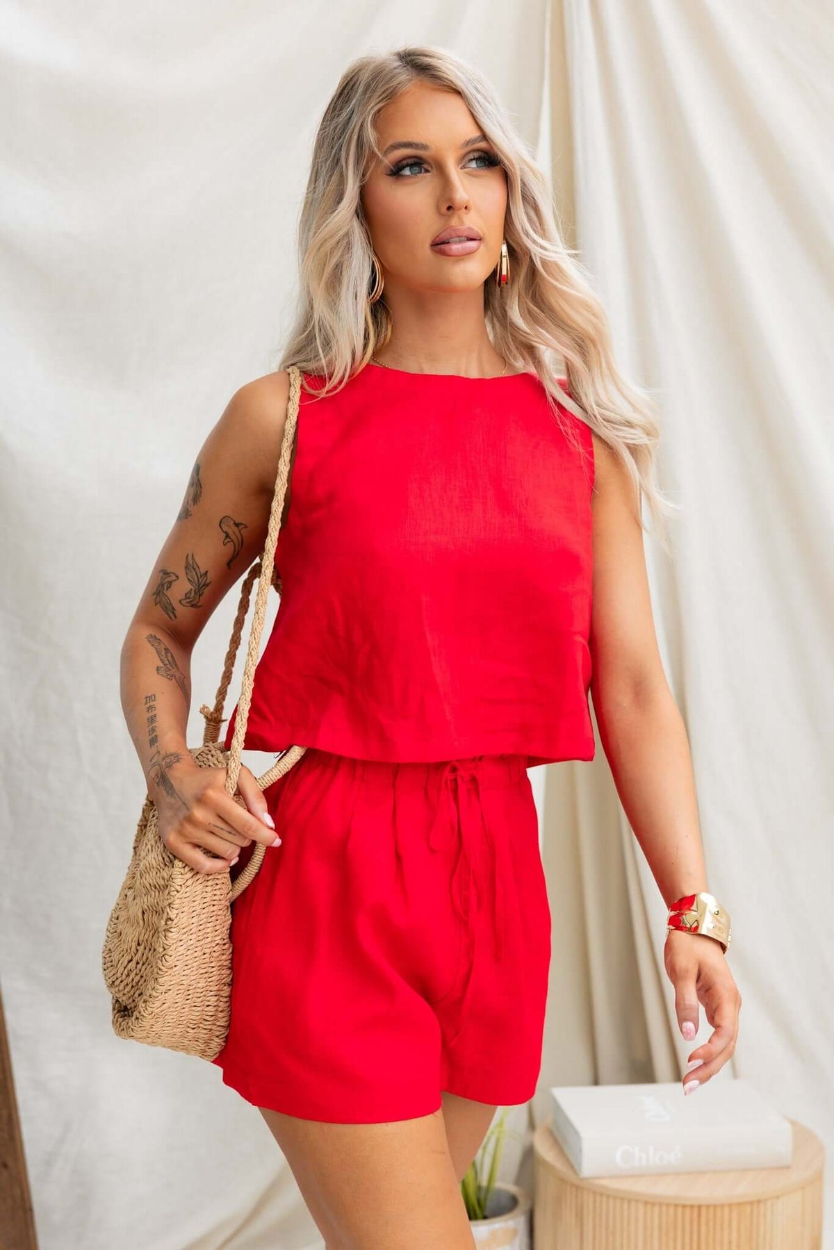 Stylish woman wearing a red linen Orson Top paired with shorts, holding a straw bag, standing in a fashion-forward pose.
