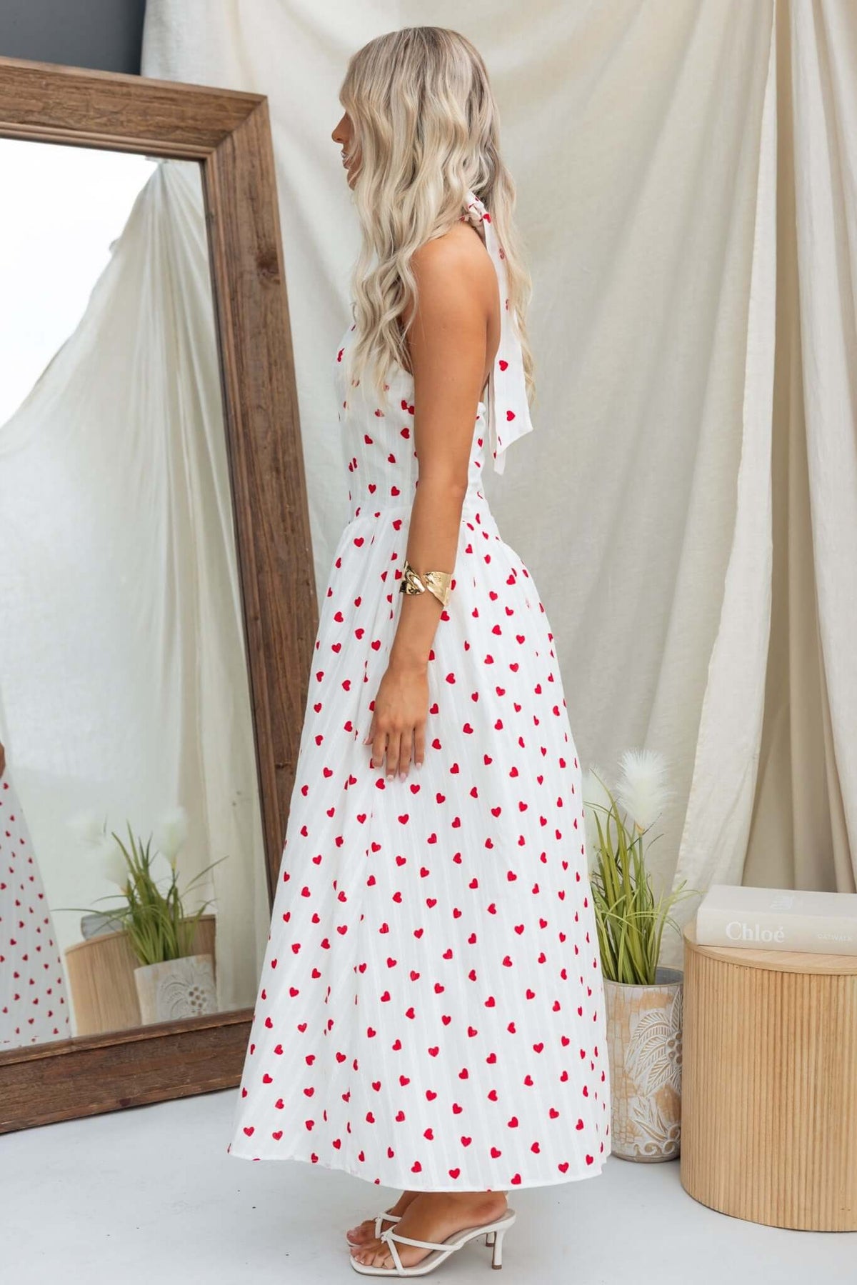 Side view of woman wearing Imarnie Maxi Dress with tie neckline and red heart print, featured in a bright, airy setting.