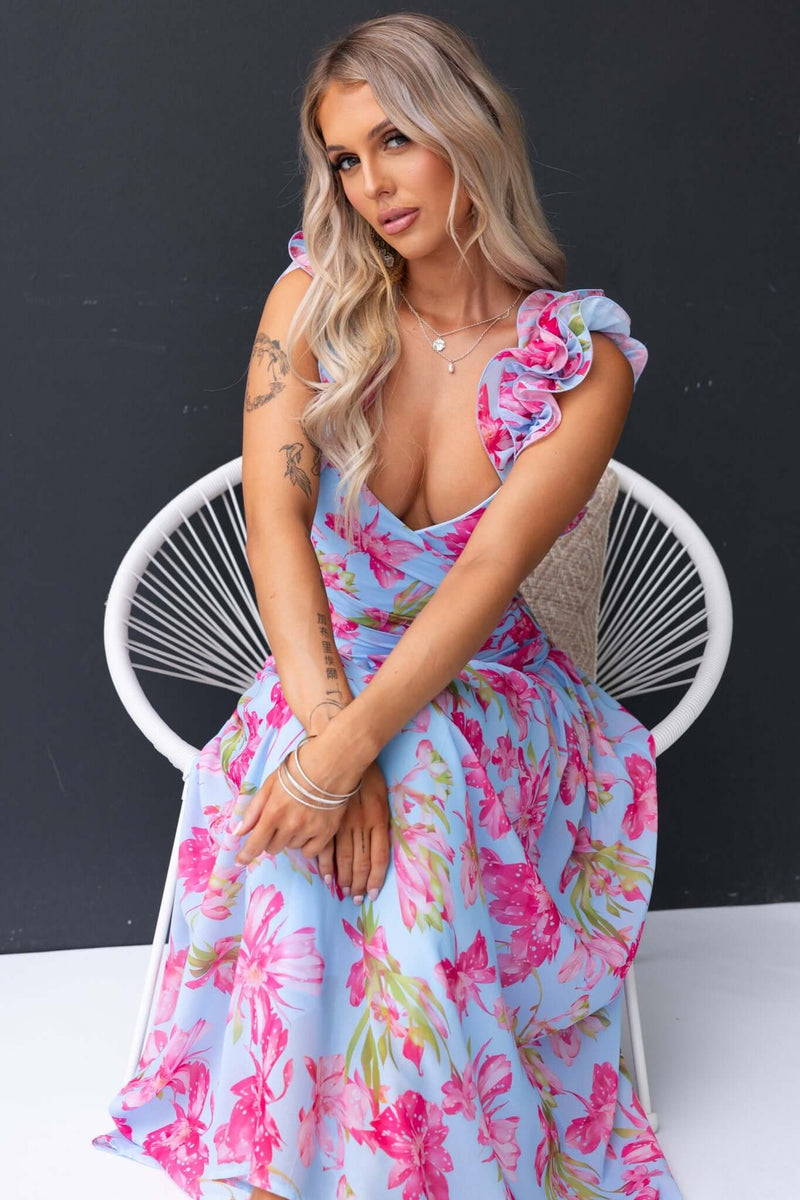 Model wearing Neleta Maxi Dress with floral design and ruffle straps, seated in a stylish chair.