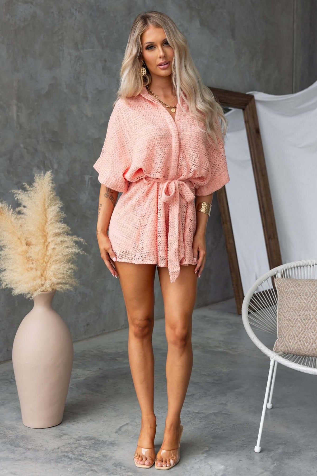 Model wearing a coral Joner Playsuit with a collar, button-up front, and removable tie, styled in a chic indoor setting.