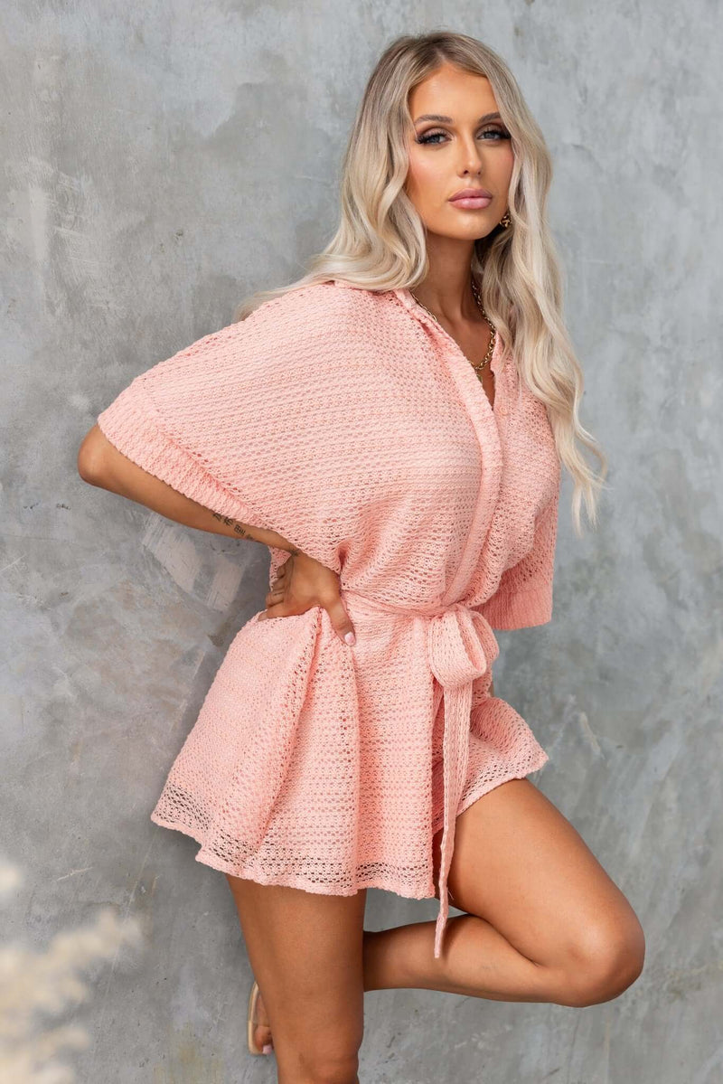 Model wearing a pink Joner Playsuit with a collar, button-up front, and tie, showcasing a relaxed fit against a textured backdrop.