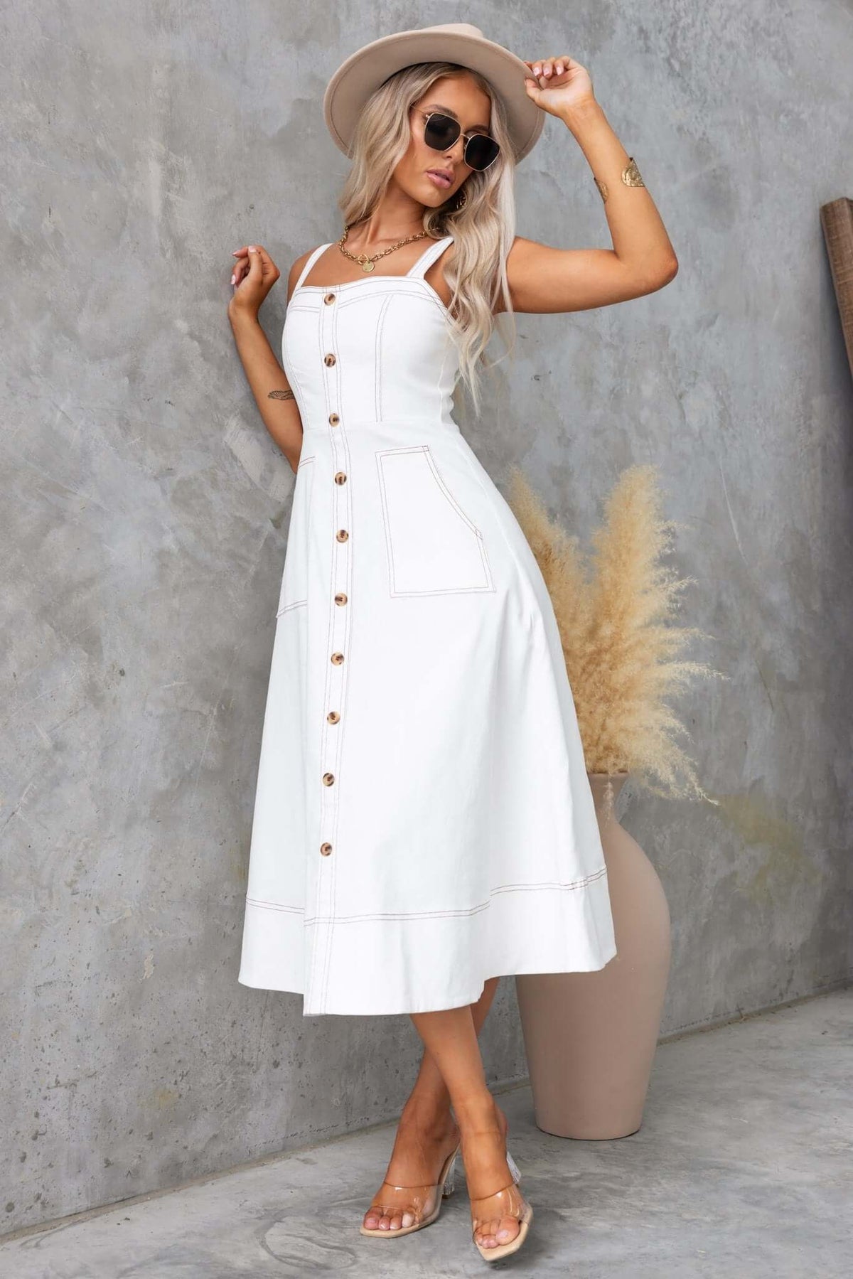 White midi dress with buttons, pockets, and spaghetti straps, styled with a hat and sunglasses against a textured wall.