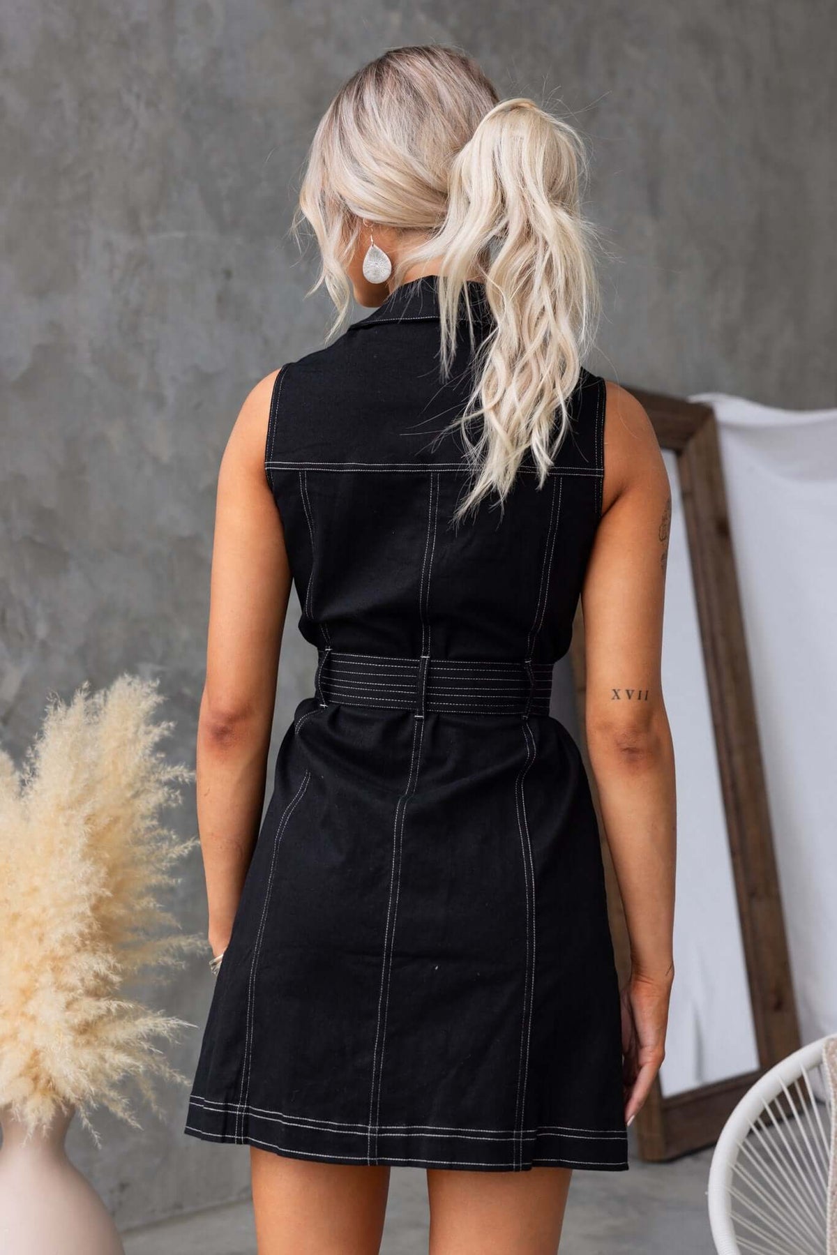 Back view of a woman in a sleeveless black denim dress with a collar and removable tie, showcasing pockets.
