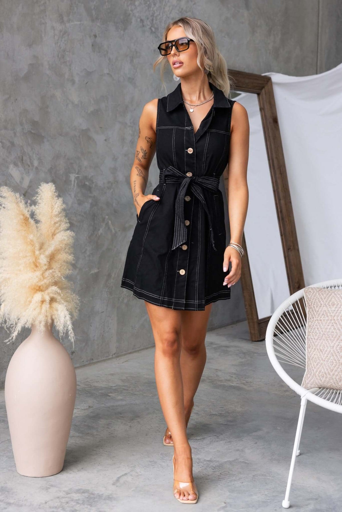 Model wearing a Tied In Denim Dress, featuring a collar, button-up front, pockets, and a removable tie, styled with sunglasses.