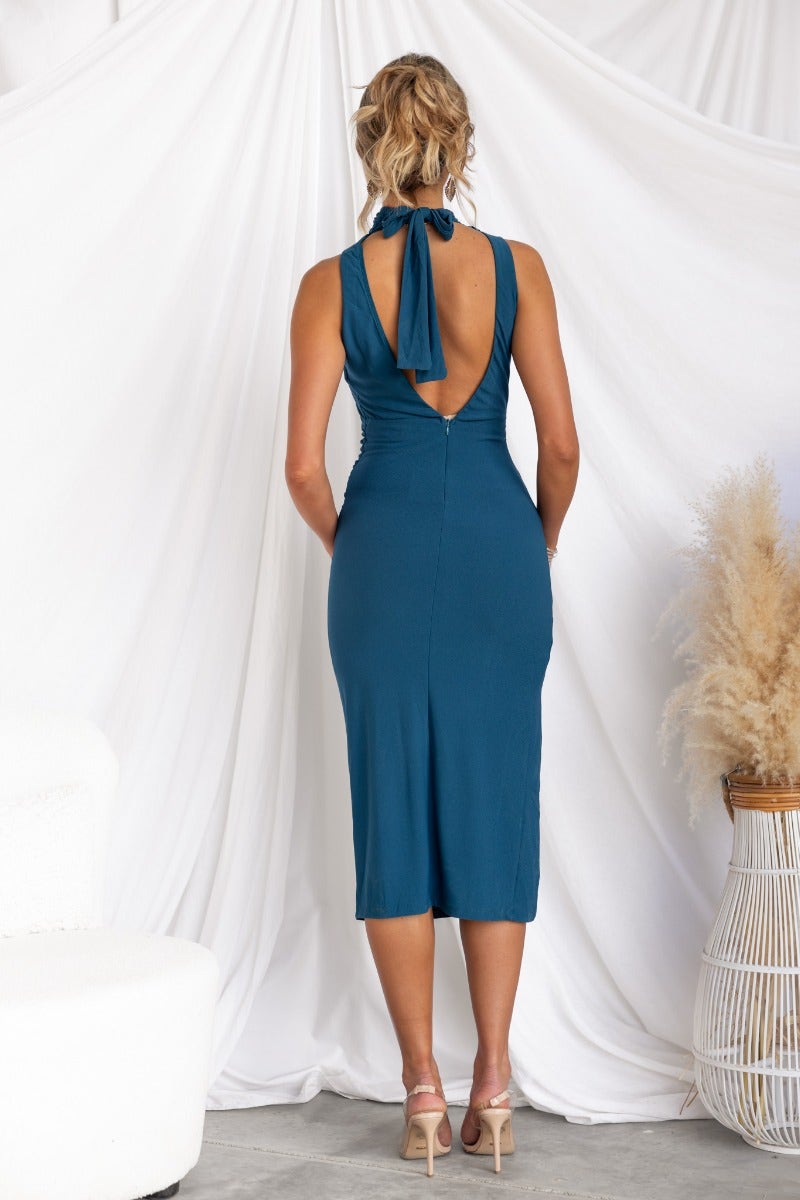 Back view of a woman in a teal bodycon midi dress with a high tie neckline and side split, styled with heels.