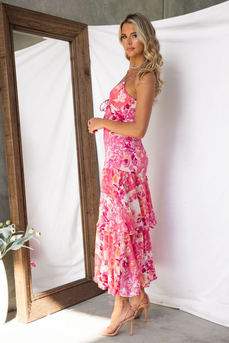 Side view of a model in a pink floral print maxi dress with adjustable straps and a tie detail at the bust.