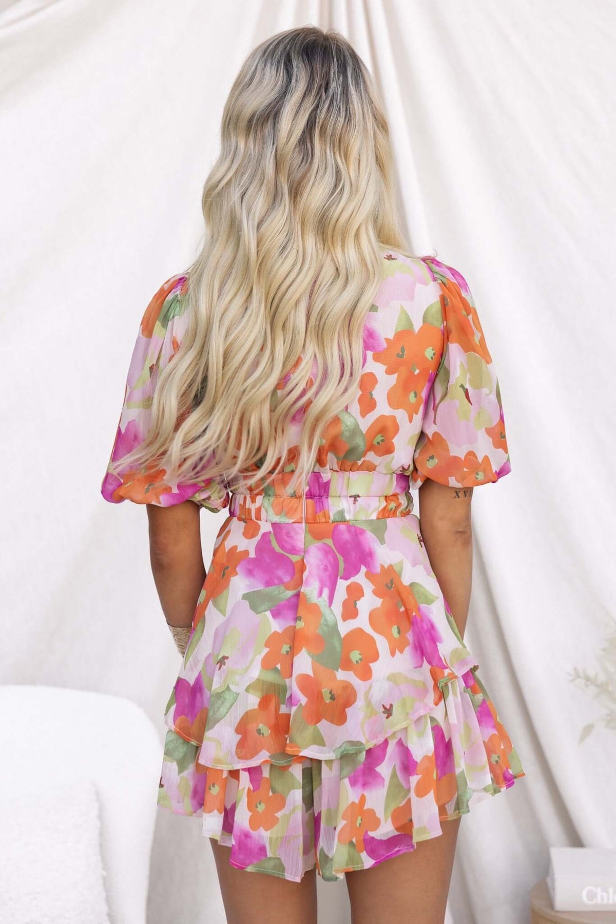 Back view of a woman wearing a floral Cyra Playsuit with ruffled skirt, showcasing vibrant colors and a fitted waist.