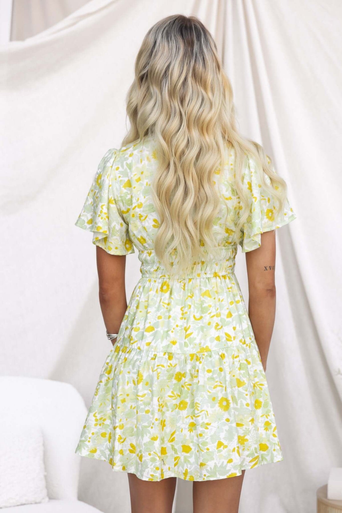 Back view of a woman in a floral Isha Dress with a V-neckline and tie at the bust, featuring flared sleeves.