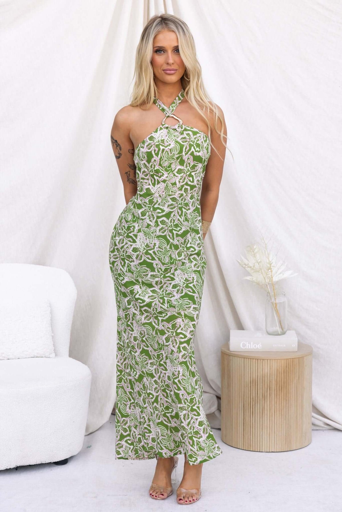 Model wearing Nino Maxi Dress with floral pattern, adjustable tie neckline, and gold hardware detail in a studio setting.