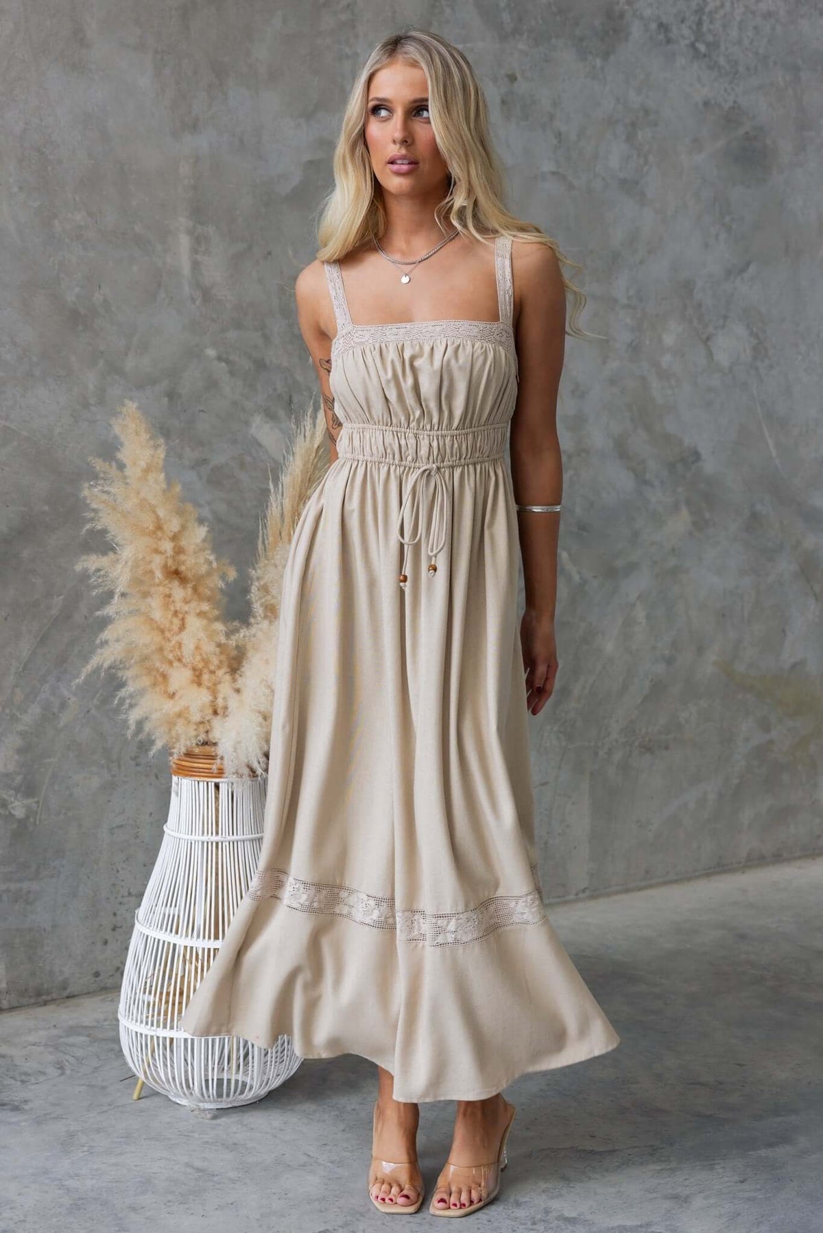 Woman wearing Eletti Midi Dress with lace detailing, elastic waist, and tie feature in a neutral color against a textured background.