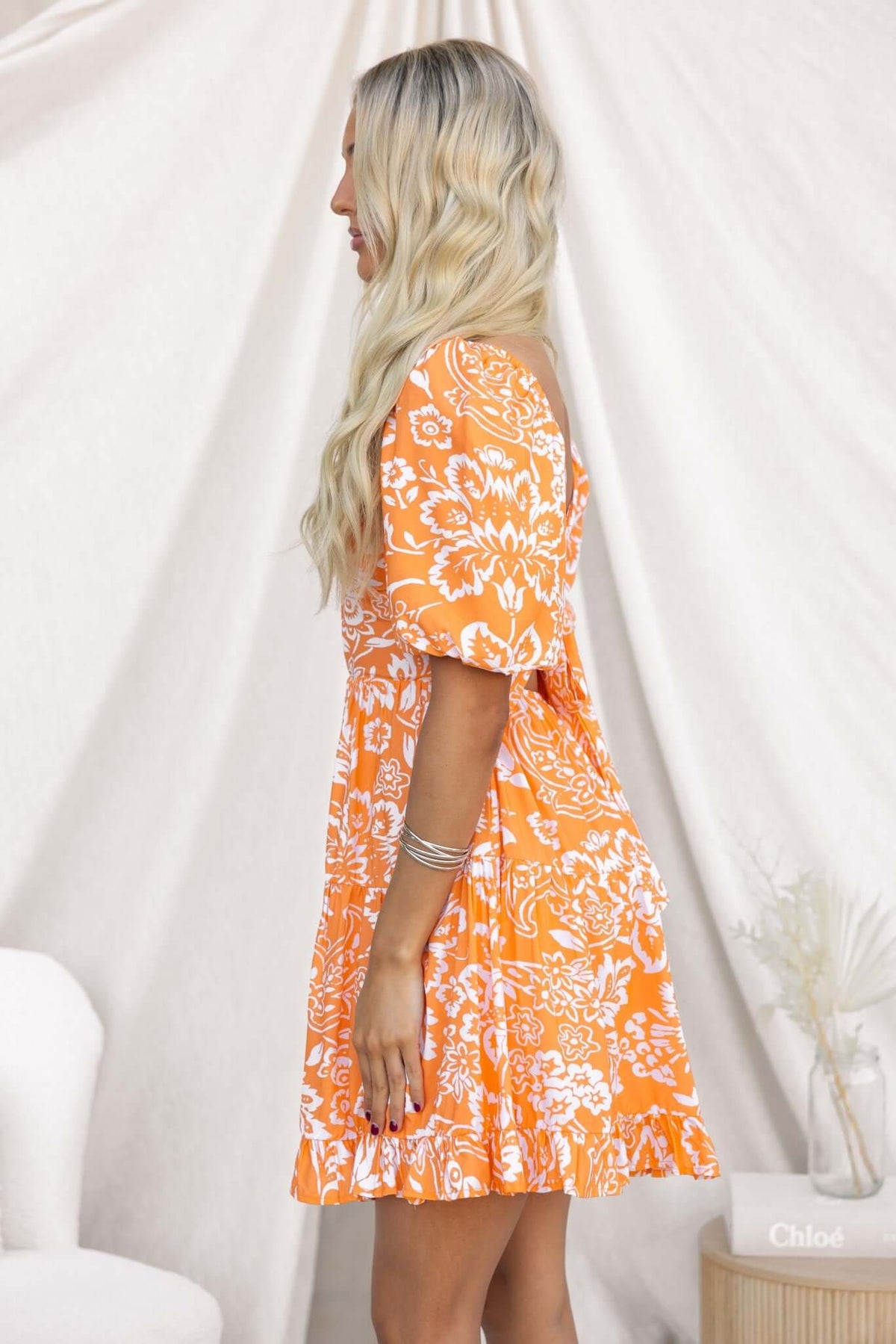 Model wearing an orange floral Omaha dress with puff sleeves, showcasing a side view in a bright setting.
