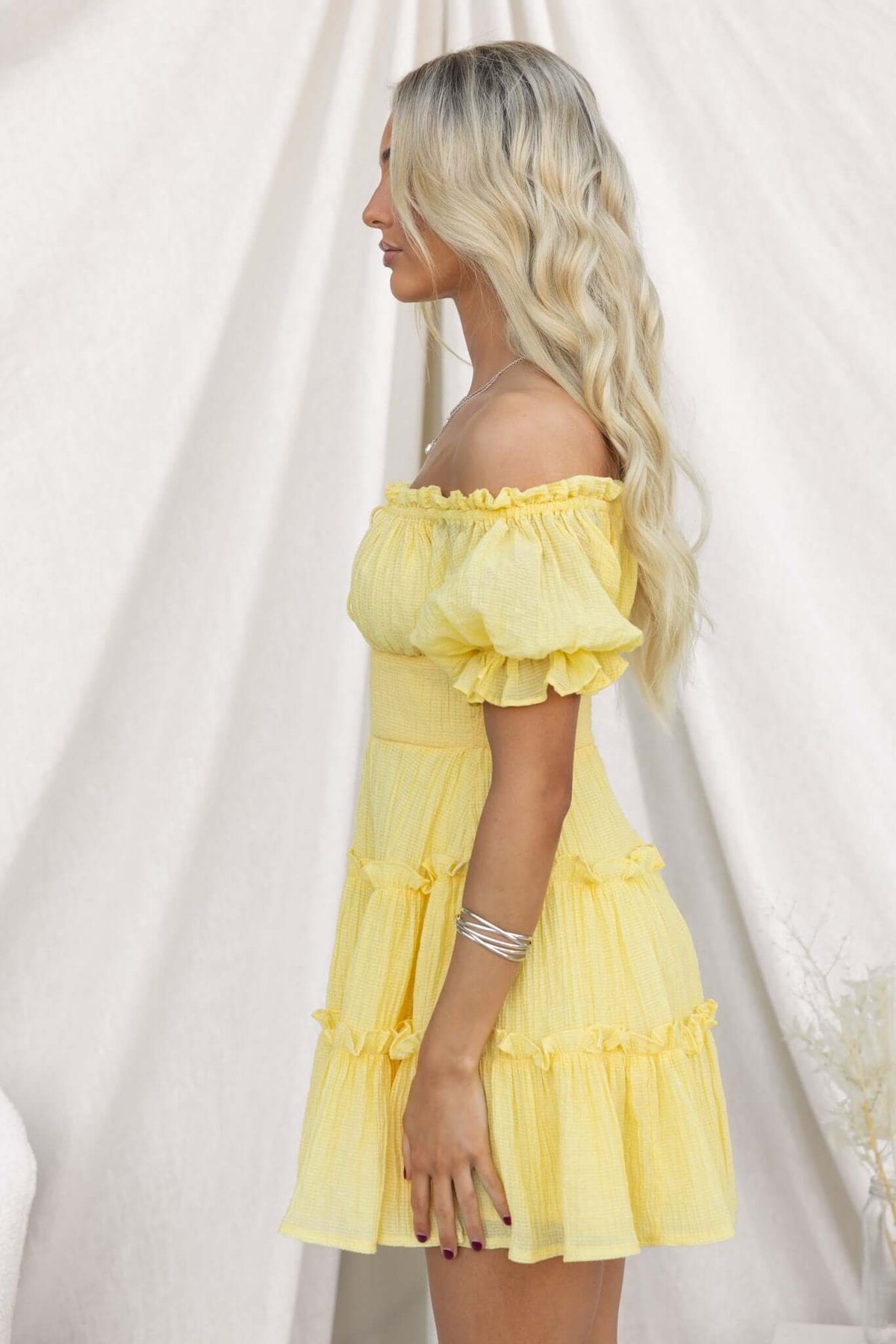 Side view of a woman in a textured yellow off-shoulder dress with tie detail and ruffle sleeves.