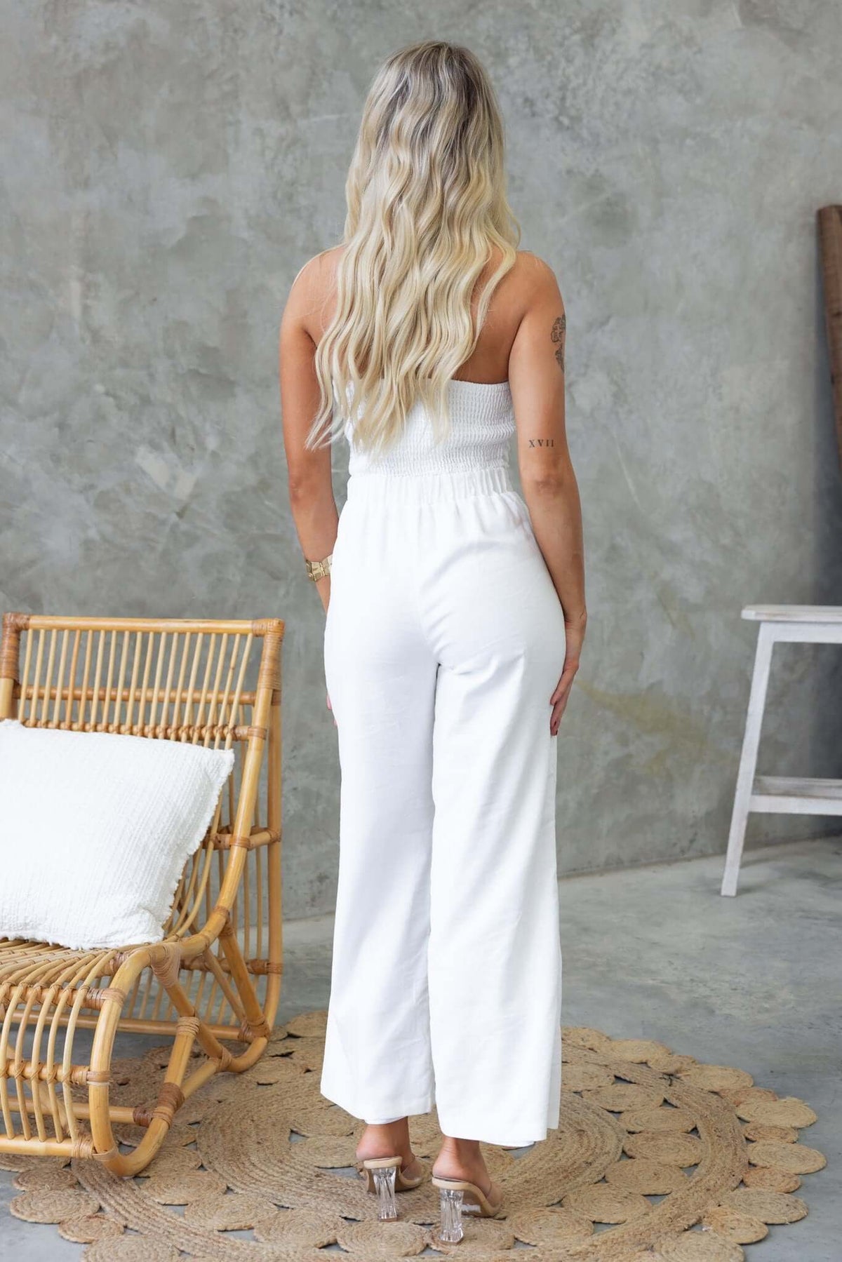 Back view of a woman in a strapless white Tuscon jumpsuit with elastic waist and pockets, styled in a modern setting.