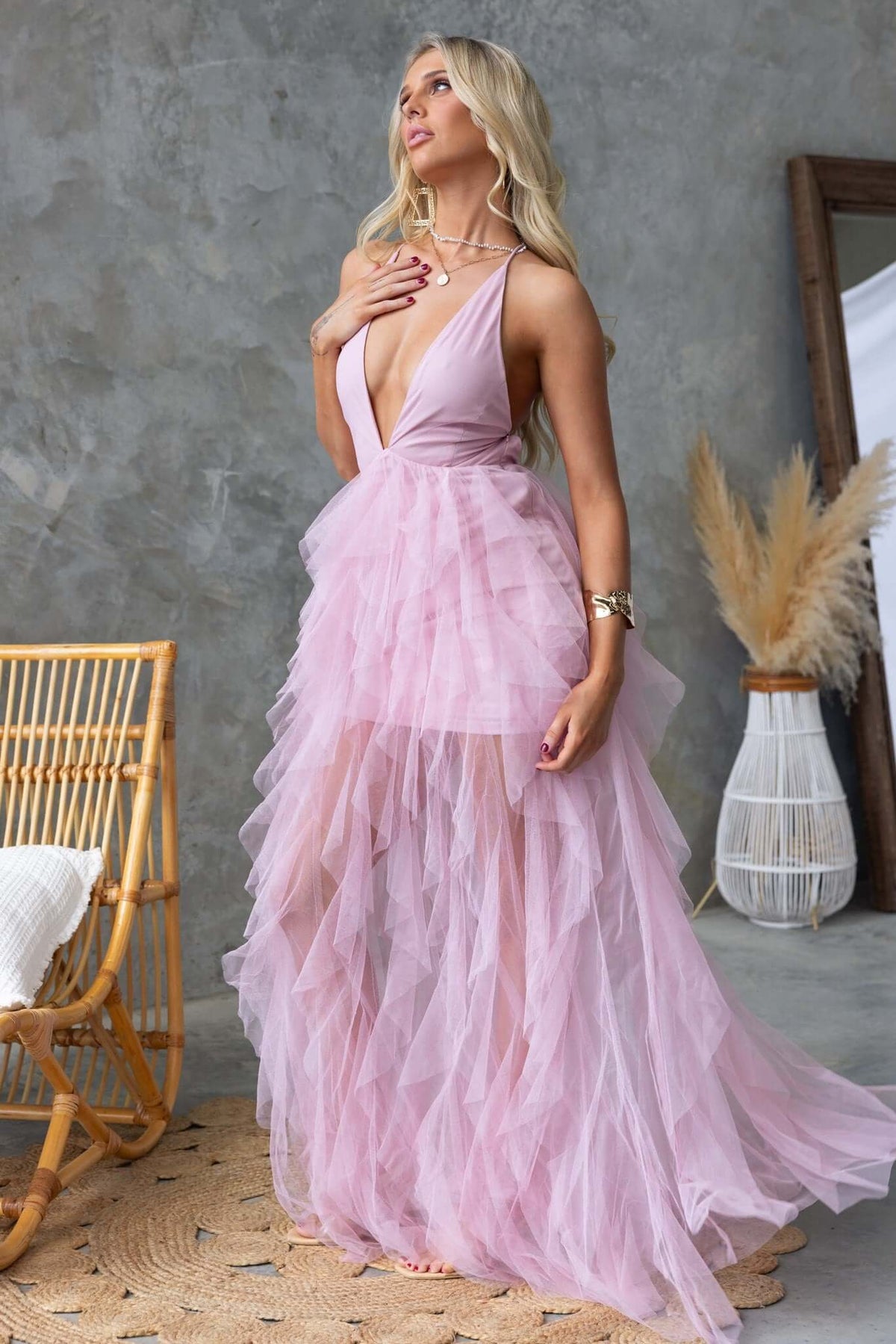 Flowy pink Salinas Maxi Dress with V-neckline, lace-up back, and tulle ruffle skirt, styled in a chic indoor setting.