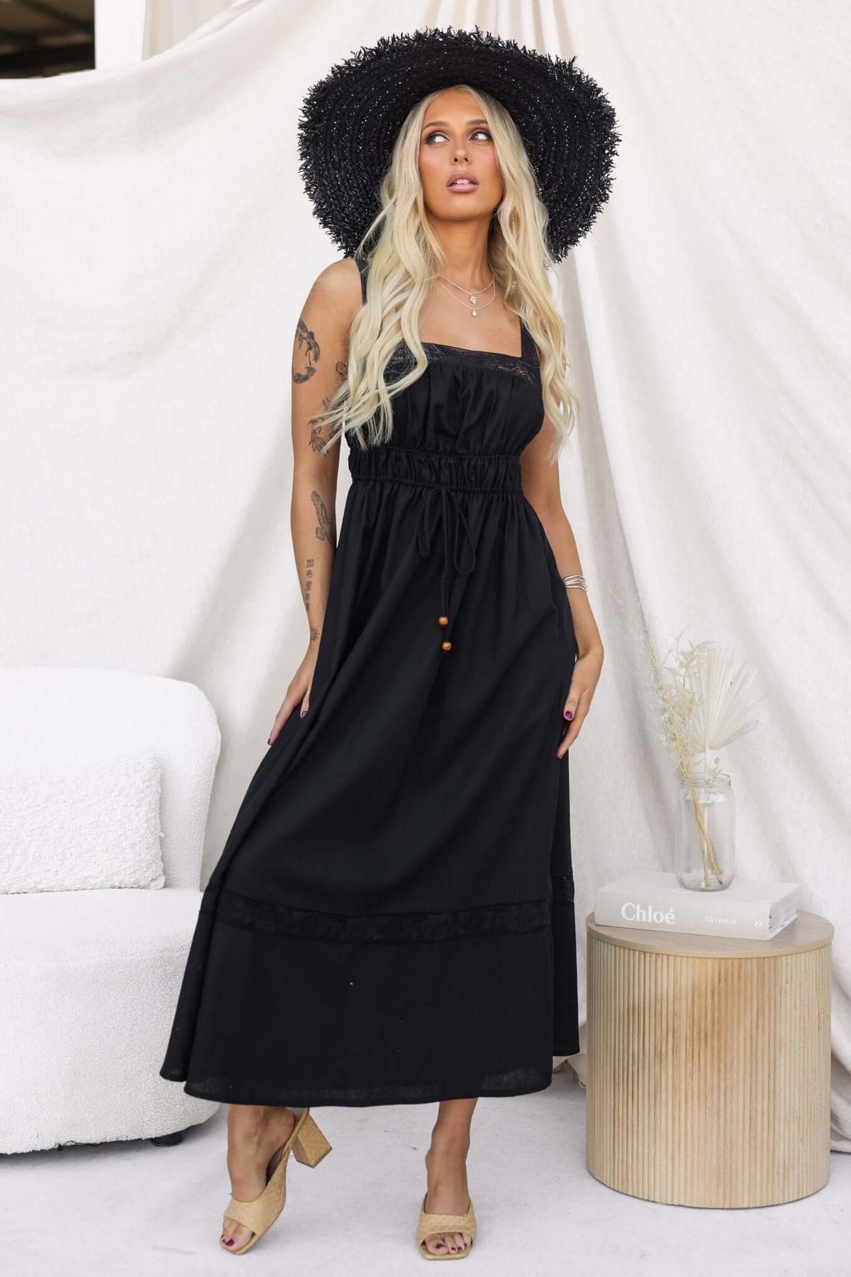 Woman wearing a black midi dress with lace detailing, elastic waist, and a large straw hat, posing indoors.