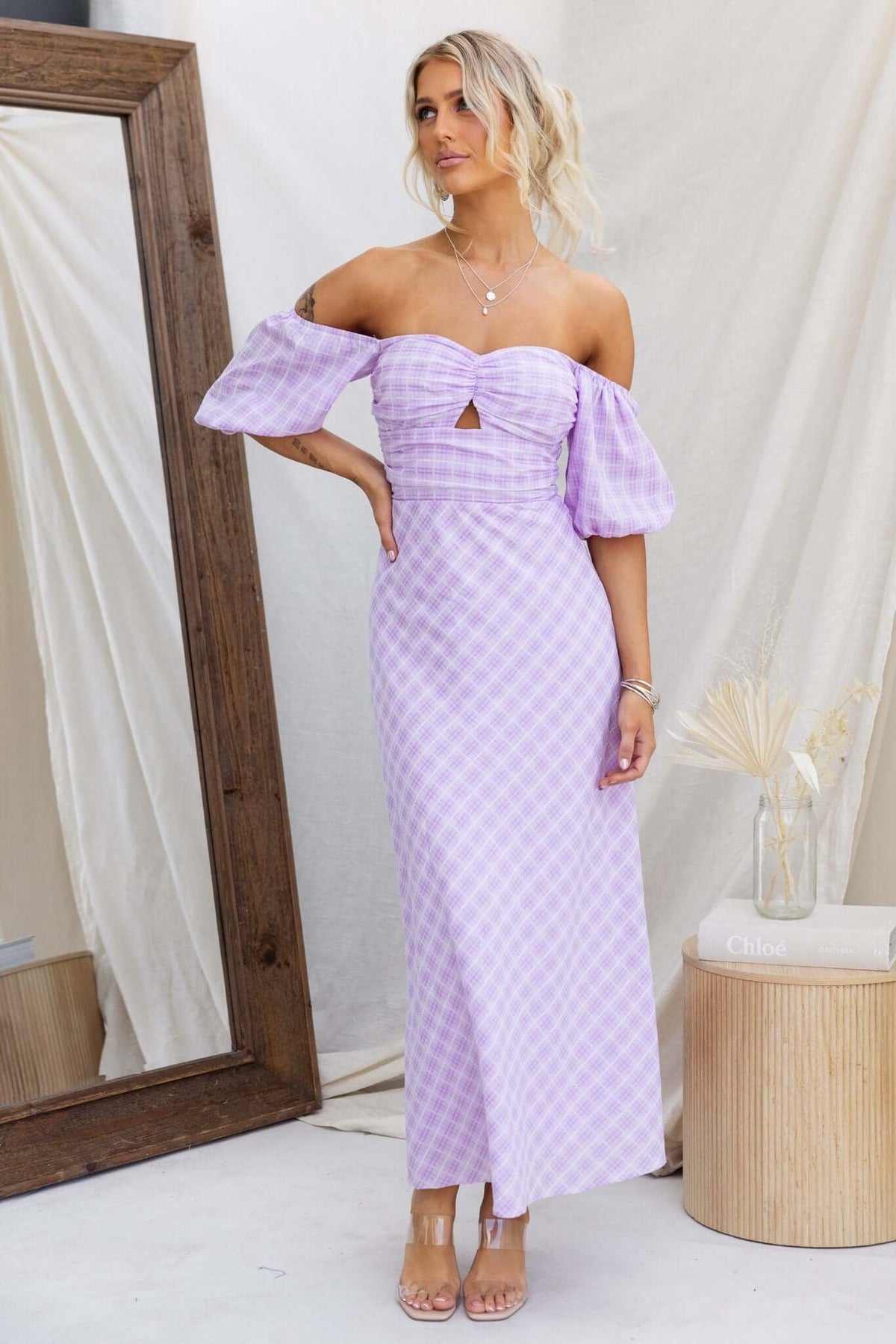 Woman in a lilac gingham Elayna maxi dress with puff sleeves and cut-out under bust, styled in a chic setting.