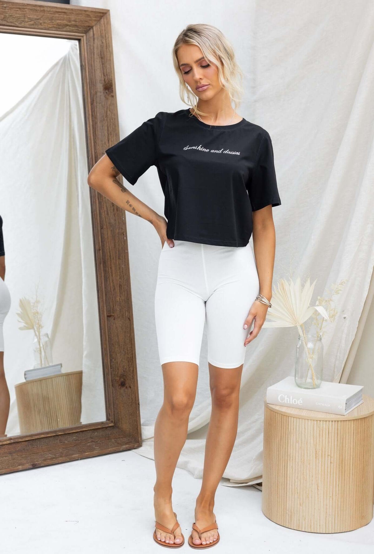 Model wearing a black Daisy Tee with round neckline, styled with white shorts and sandals, in a chic indoor setting.