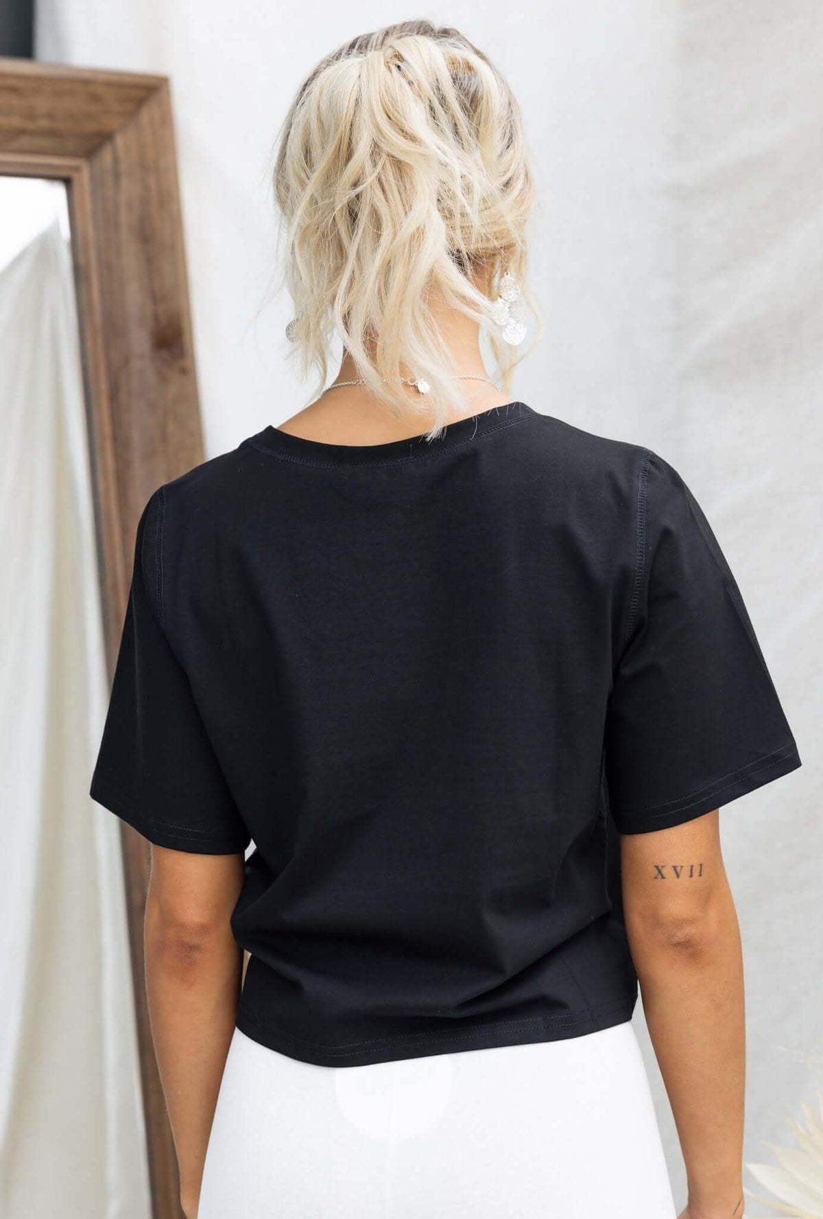 Back view of a woman wearing a black Daisy Tee with round neckline and short sleeves, styled with white bottoms.