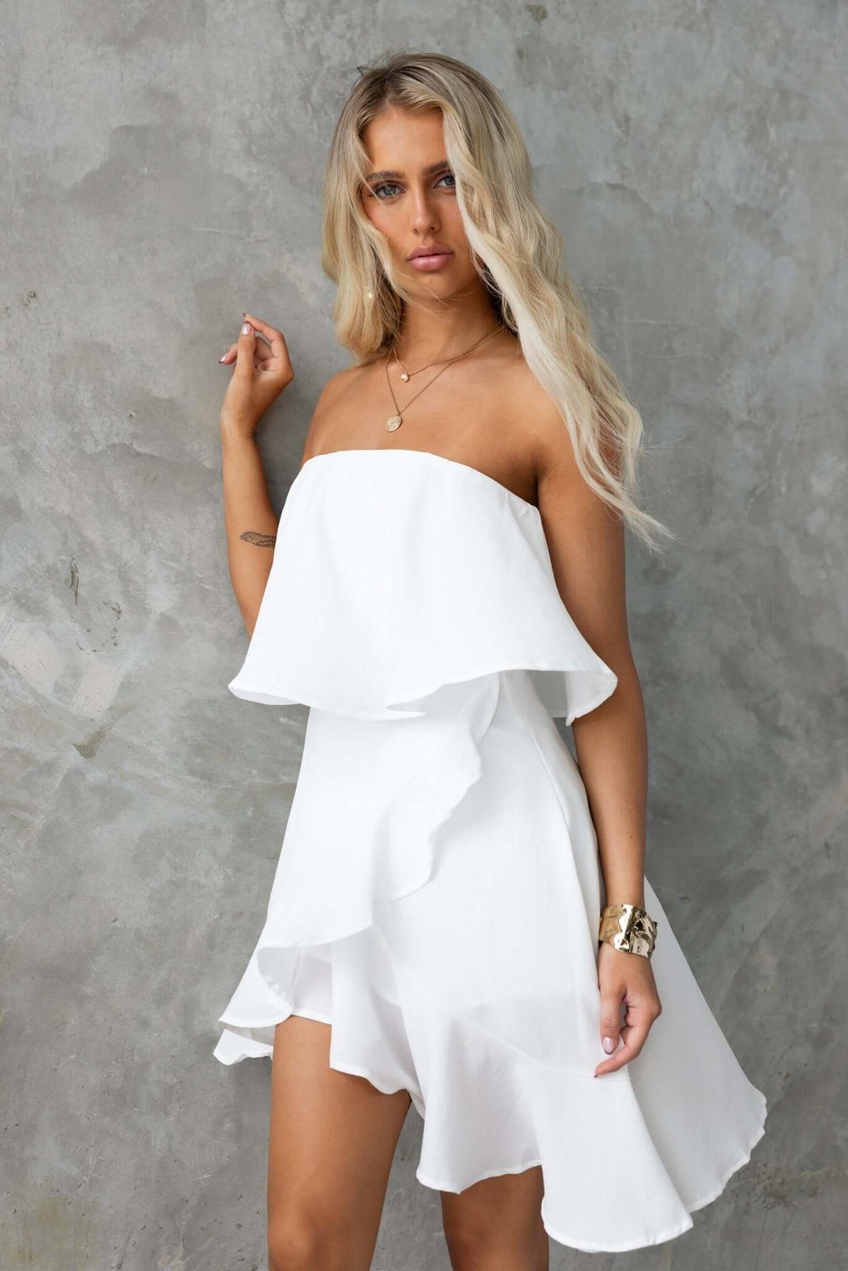 Model wearing a white strapless Adee dress with ruffled layers against a textured grey background.