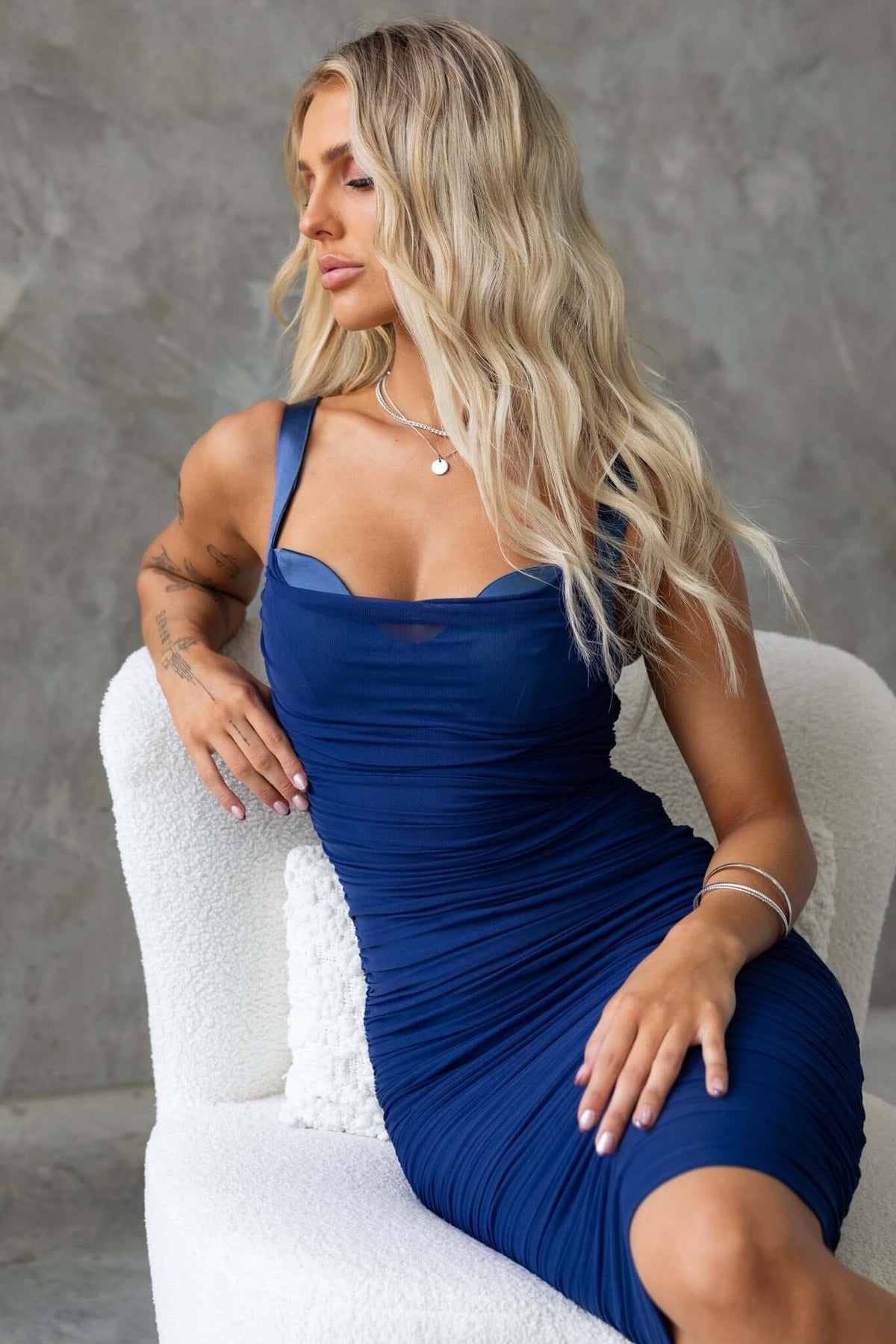 Stylish model wearing a fitted blue midi dress with adjustable straps and ruched design, seated elegantly.