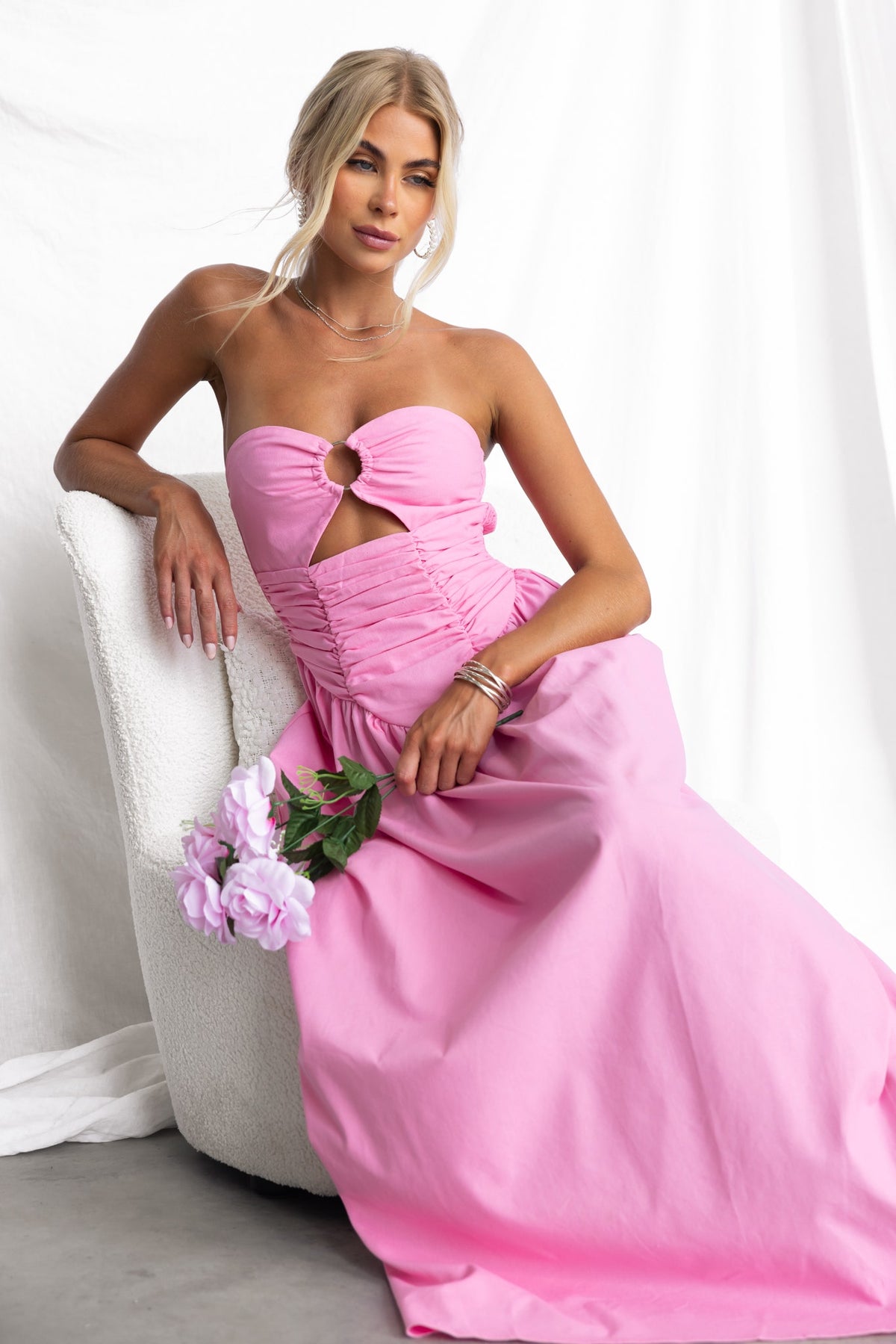 Model wearing a pink Abelle Maxi Dress with strapless design and ring detail, seated on a chair with flowers.