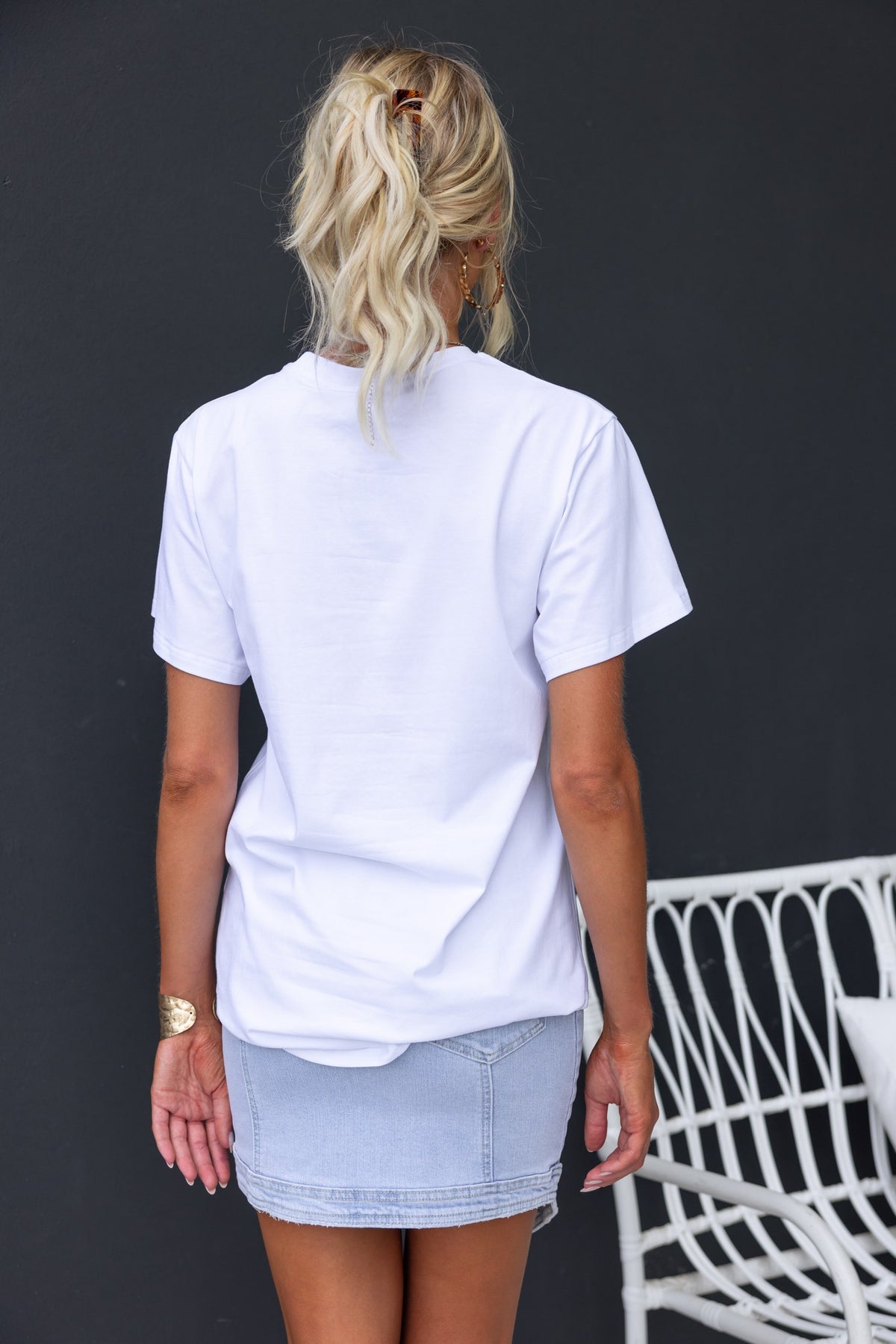 Back view of a woman wearing a white Cesca Tee paired with a denim skirt against a dark background.