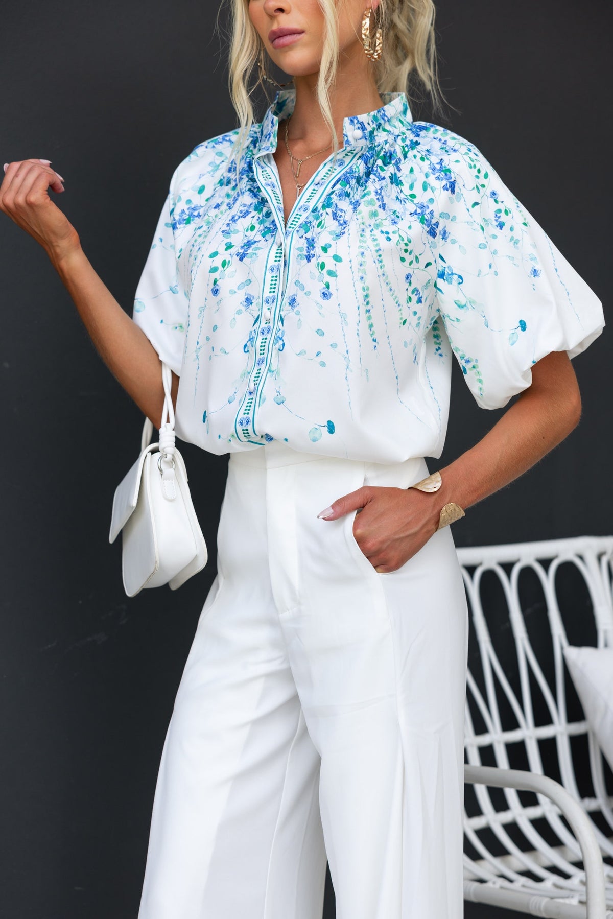 Model wearing Bruna Shirt, a floral print shirt with high neckline and button-up front, paired with white pants.