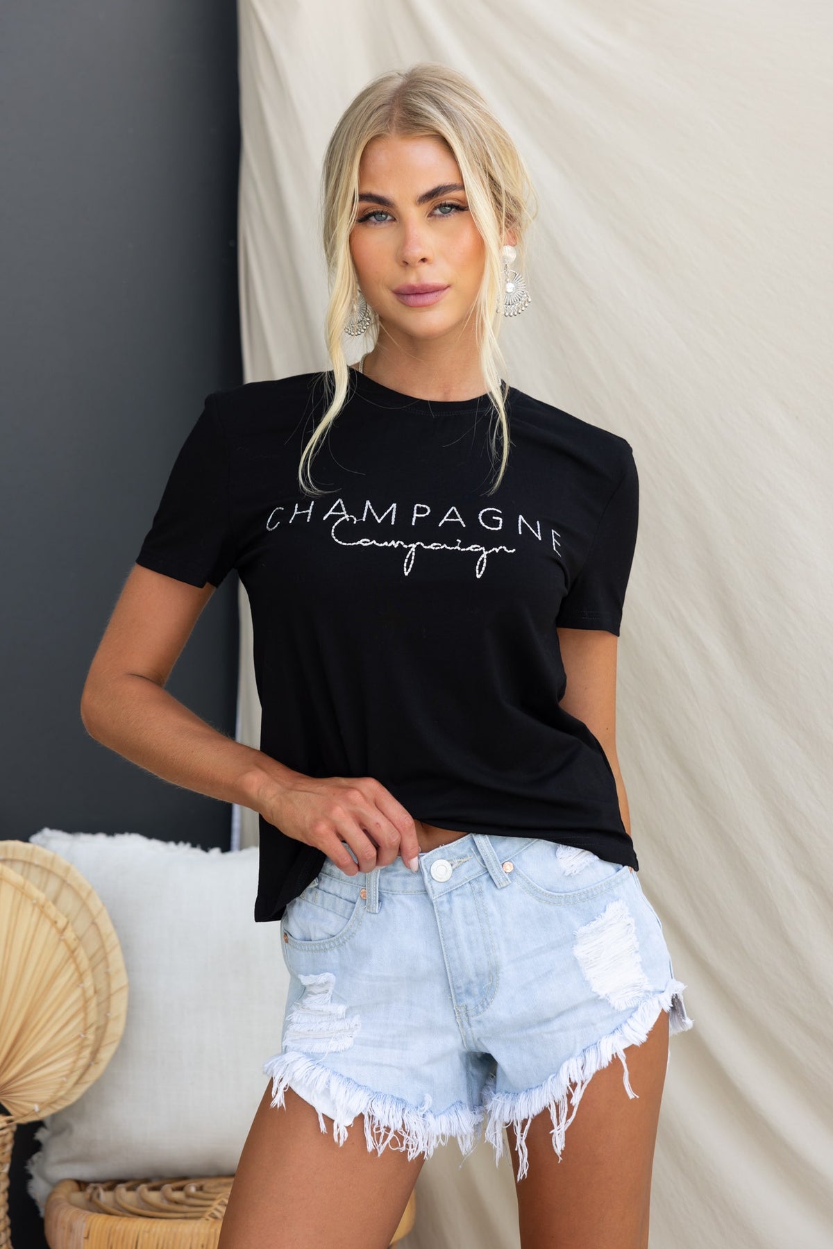 Model wearing black Wyatt Tee with "Champagne campaign" text, styled with denim shorts in a bright setting.