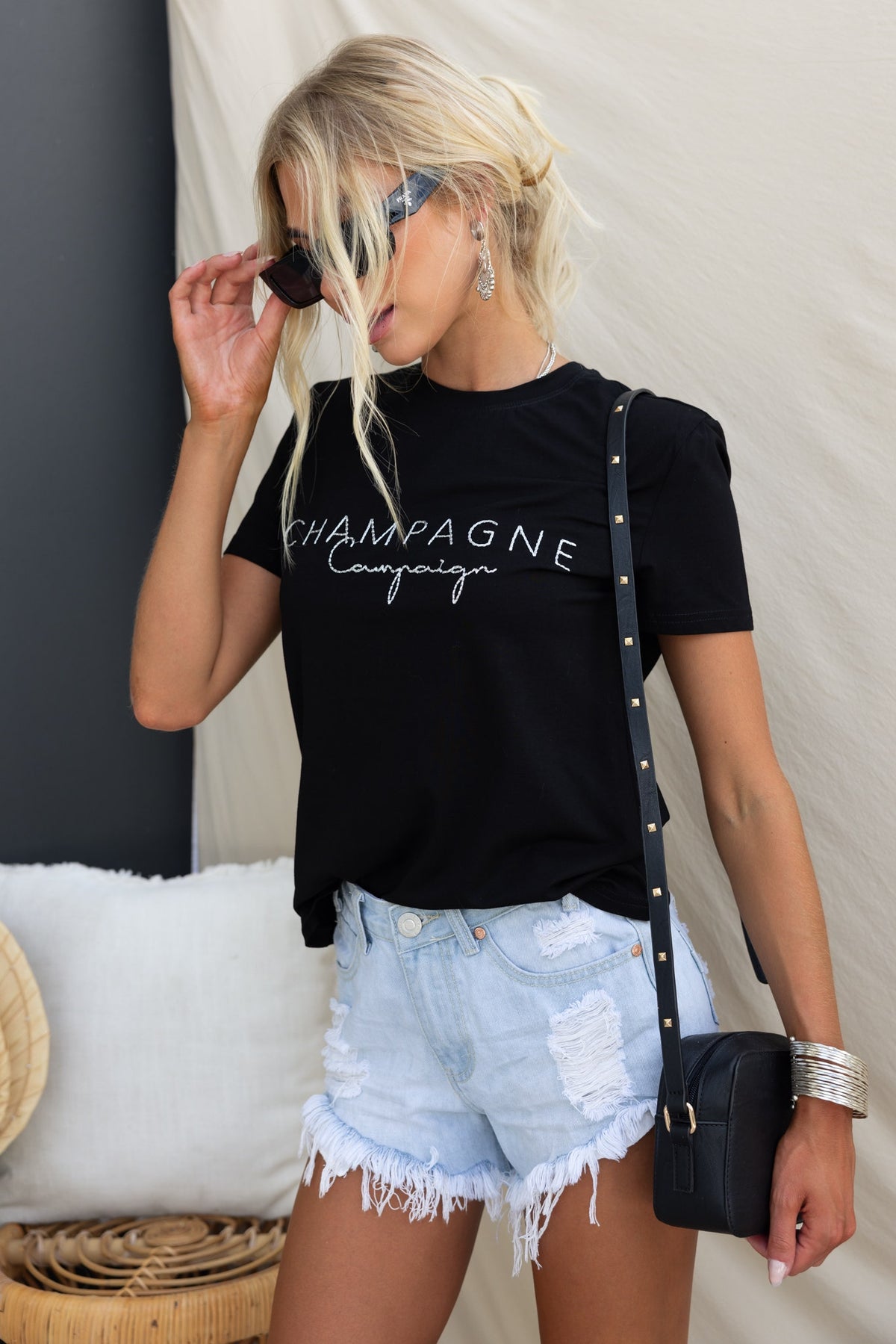 Model wearing black 'Champagne Campaign' tee with distressed shorts and sunglasses, showcasing a relaxed summer look.