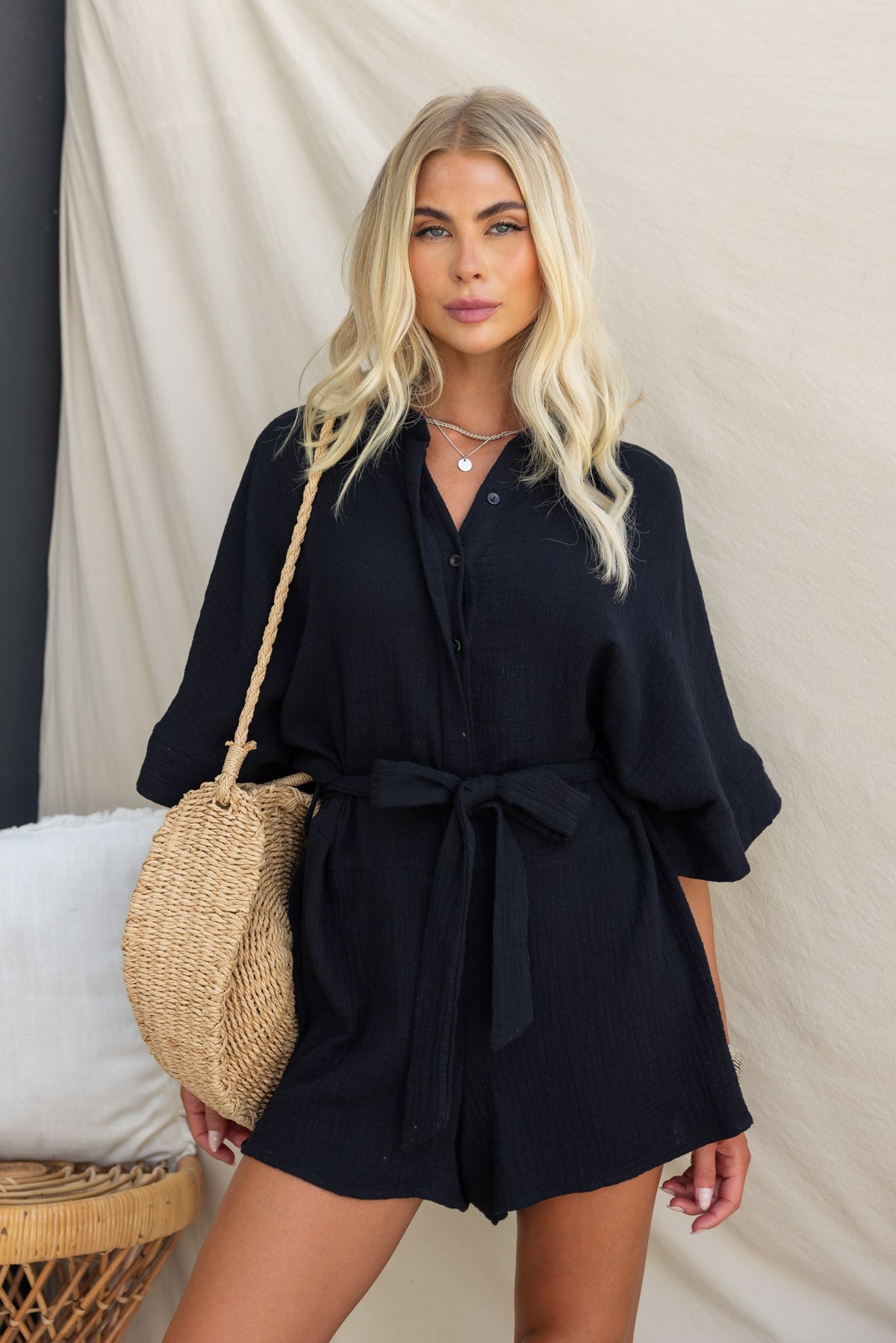 Woman in a black collar button-up playsuit with removable tie, accessorized with a straw bag.