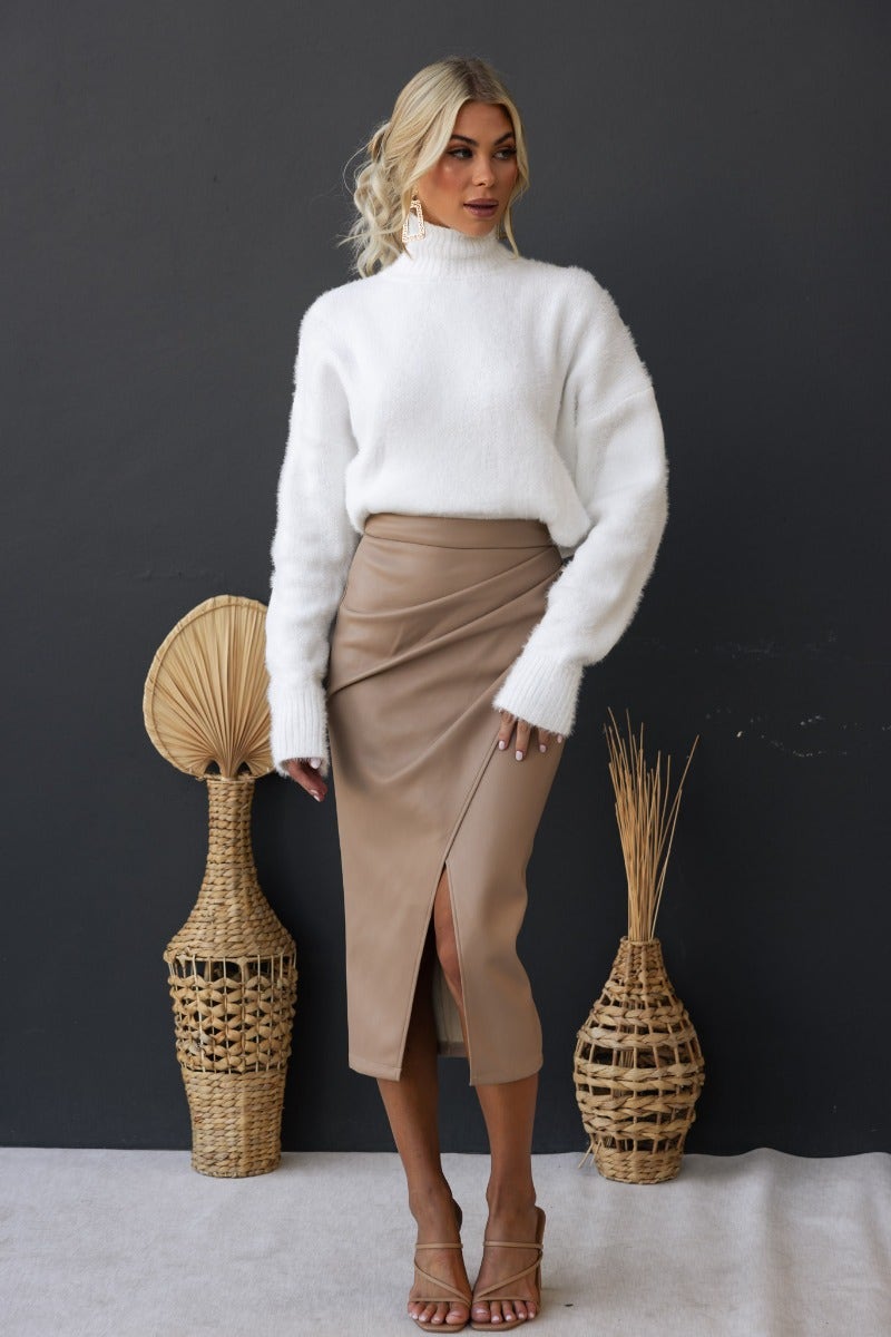 Model wearing Qorinda Midi Skirt in faux leather with asymmetrical hemline and white sweater against a dark background.