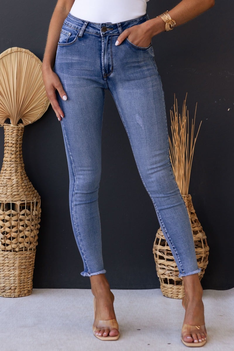 Woman wearing Juicebox Jeans with mid waist fit, distressed details, and raw hem against a dark background.