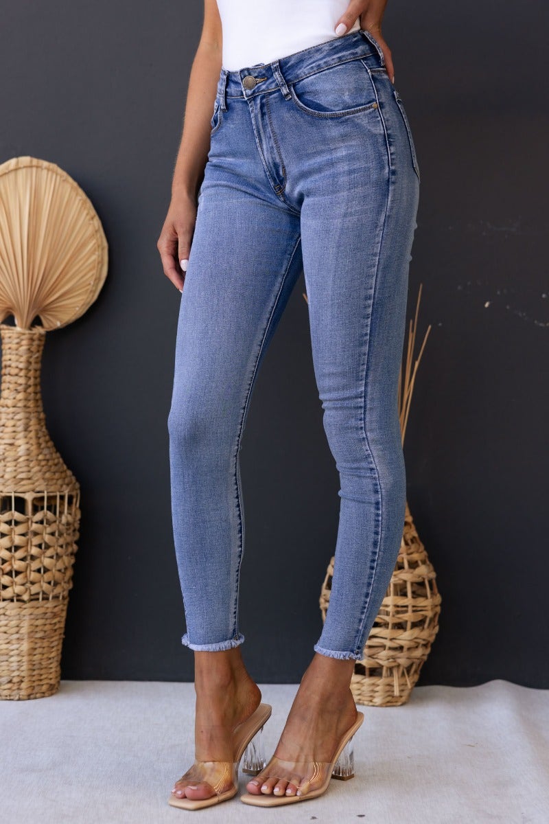 Juicebox Jeans featuring mid waist fit, distressed details, and raw hem with a standard skinny fit in a stylish light blue wash.