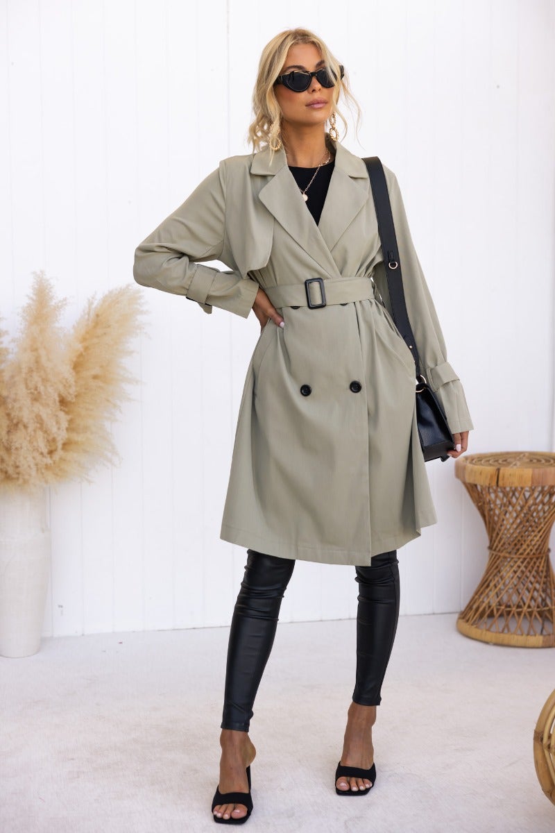 Fashion model in a stylish Cheyla trench coat with belt and sunglasses, paired with black leggings and heels.