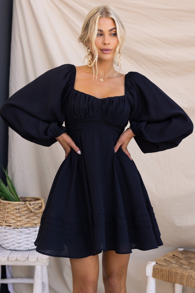 Model wearing a black Amos dress with sweetheart neckline and puffed sleeves, styled against a neutral backdrop.