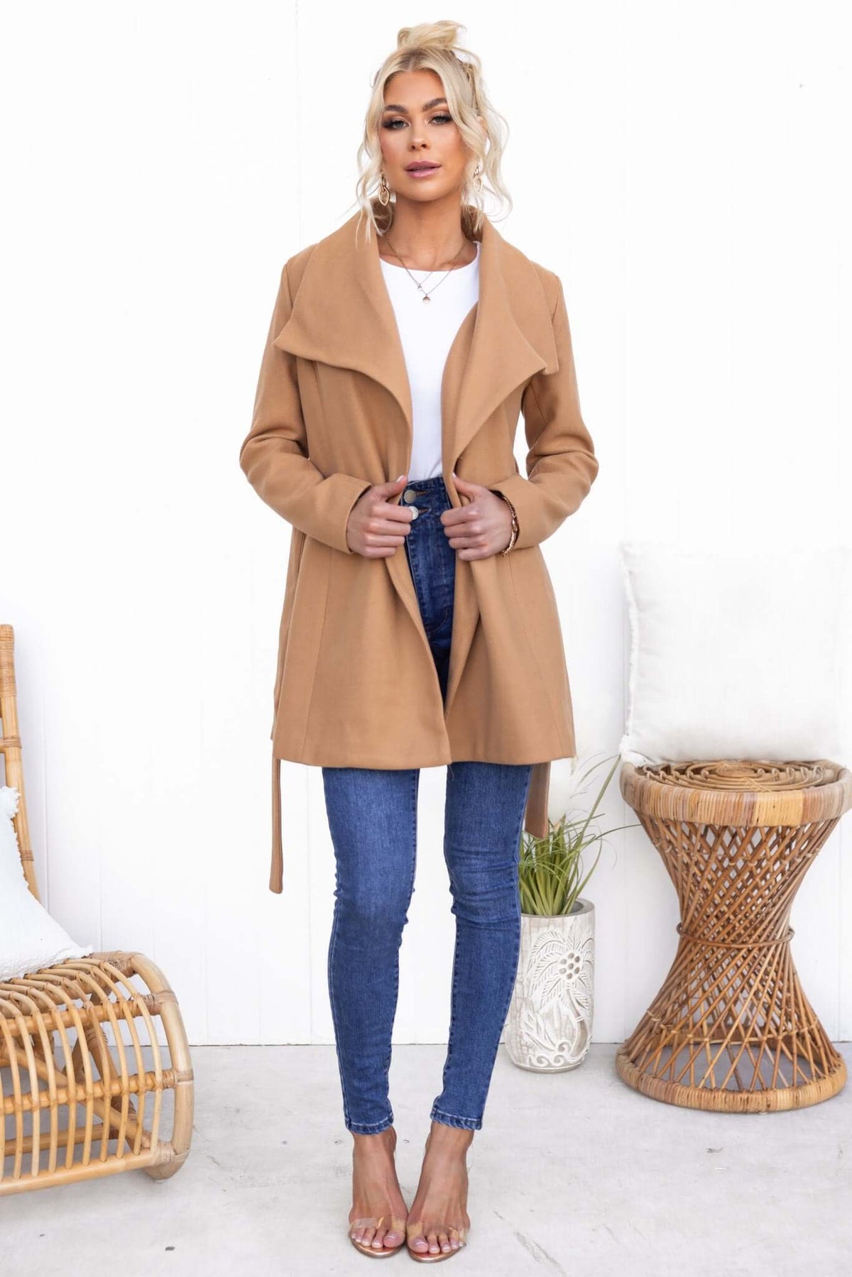 Woman wearing a stylish camel Jean Coat with a removable tie, paired with denim jeans and heels, standing against a white backdrop.