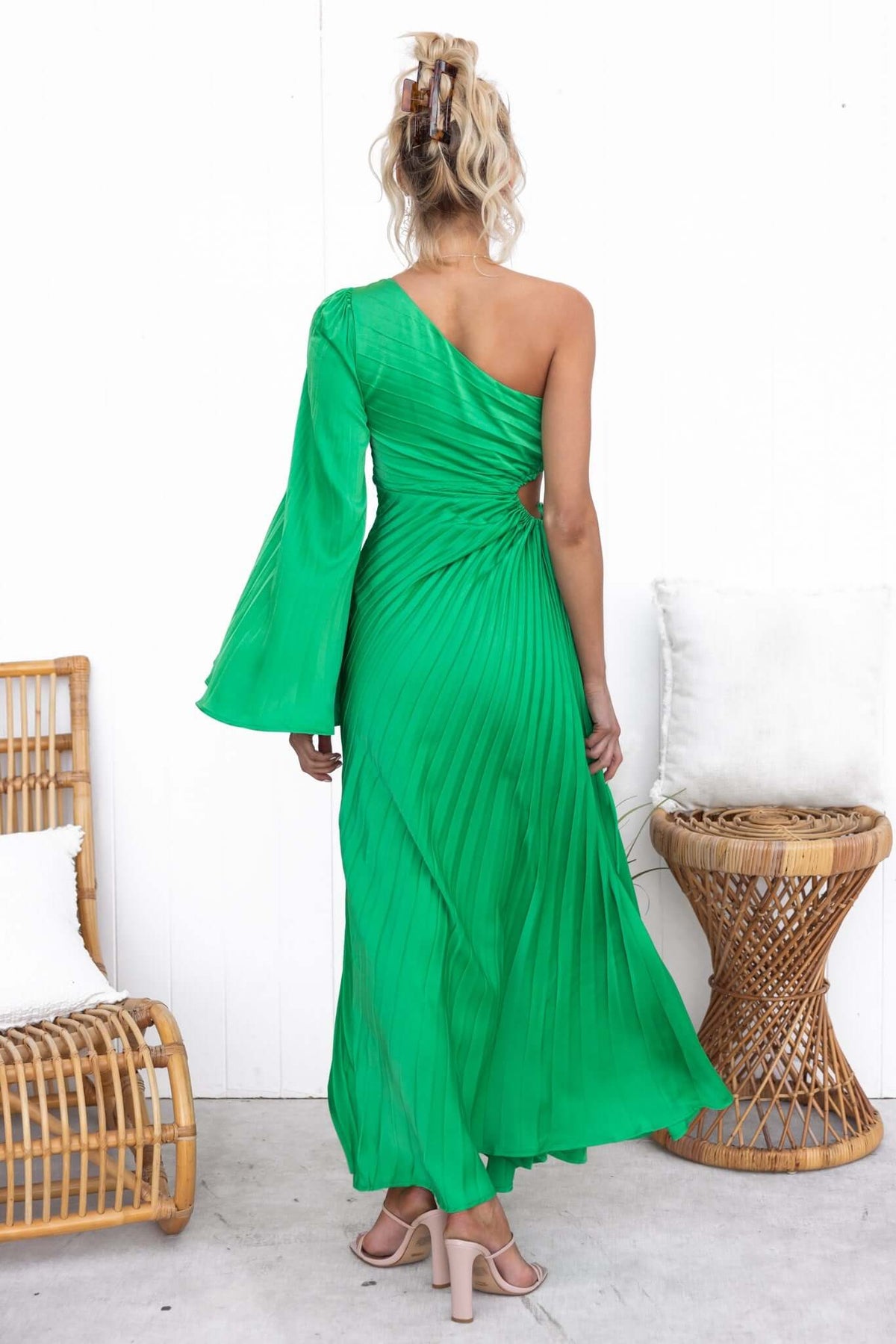 Back view of a woman in a green pleated one-shoulder maxi dress with a cut-out waist and flowing sleeves.