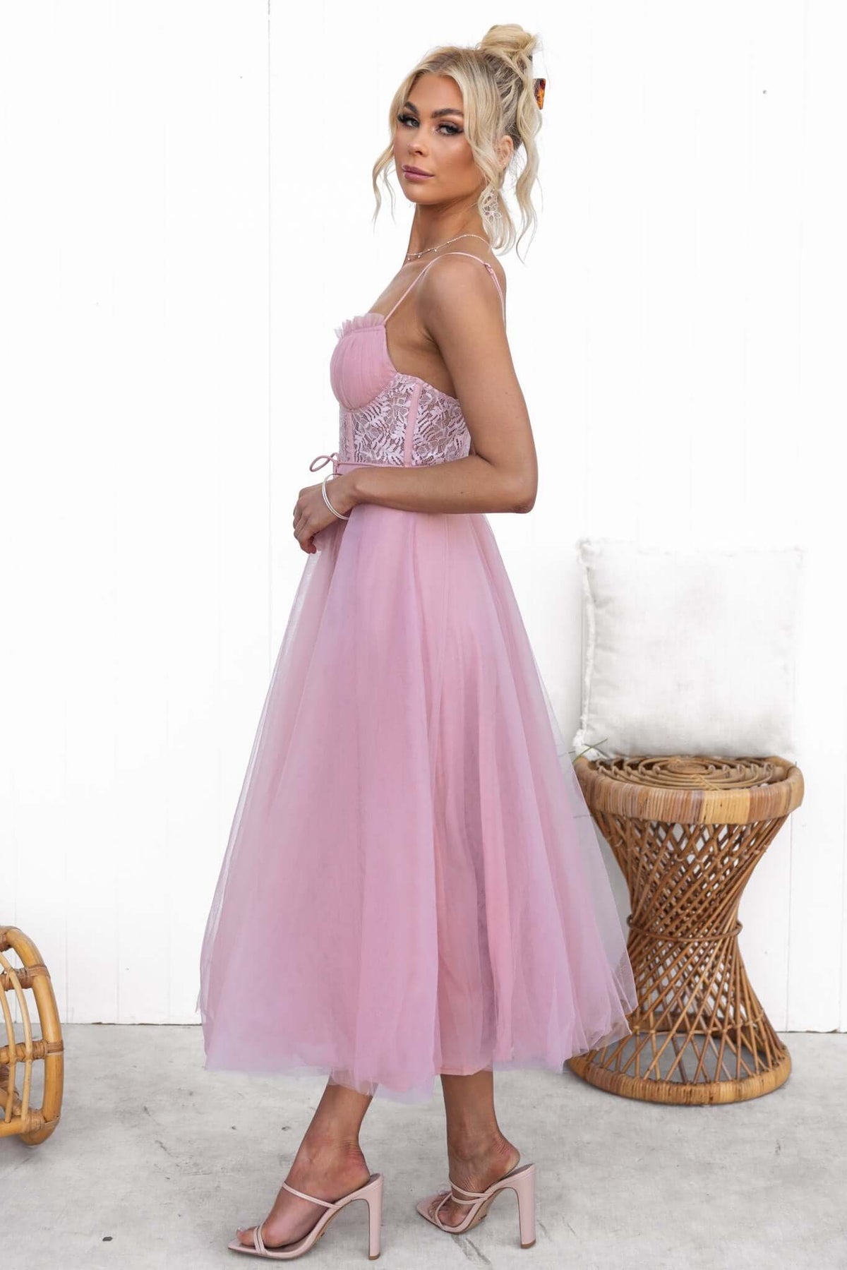Model wearing Wash Away Maxi Dress in pink tulle with adjustable straps and lace detail, standing beside a wicker chair.