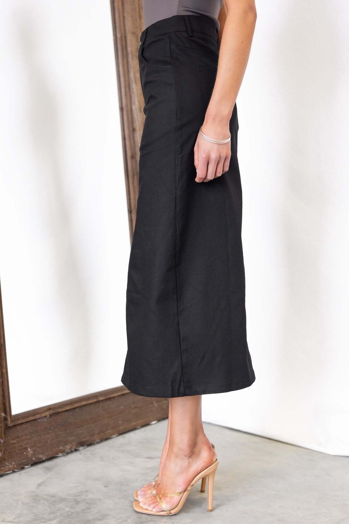 Burbank Midi Skirt in black, featuring a mid-high waist, front split, and pockets. Model wearing with heels.