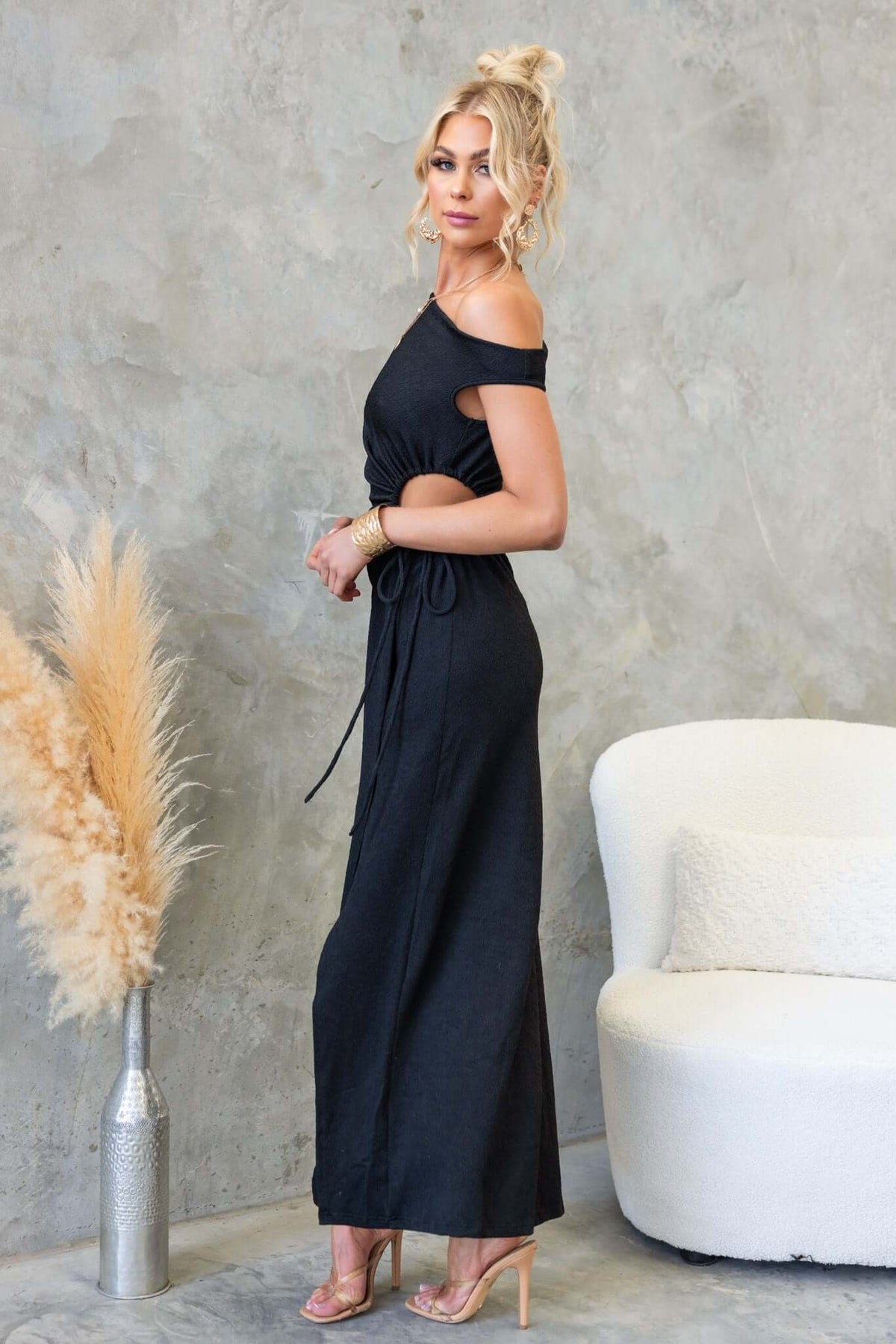 Romney Maxi Dress featuring a black asymmetric cut with tie side, model standing elegantly against a textured background.
