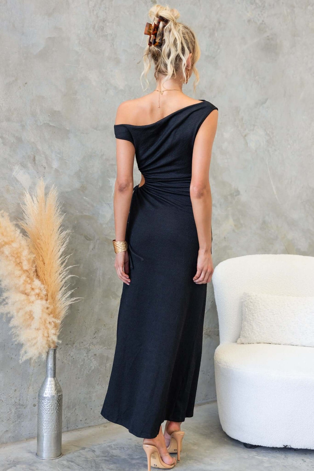 Back view of a woman wearing a black maxi dress with an asymmetric neckline and side cut-out, styled with a hair clip.