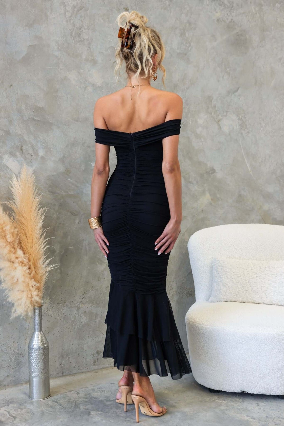 Back view of a woman in a black off-shoulder ruched maxi dress, showcasing ruffled hem and elegant fit.