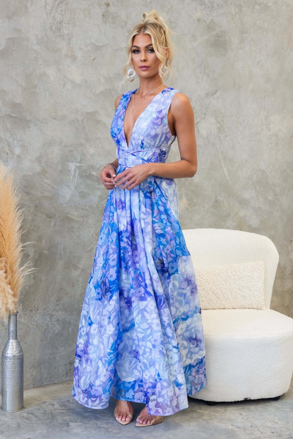 Kamala Maxi Dress in blue floral print featuring V-neckline and asymmetric hemline, styled with elegant accessories.
