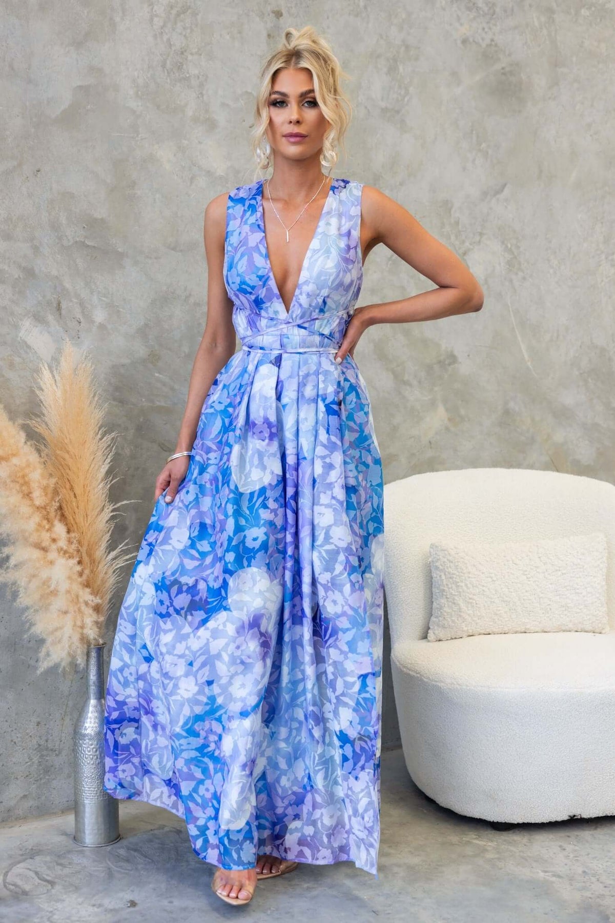 Kamala Maxi Dress in blue floral pattern with V-neckline and asymmetric hemline, styled in a modern room setting.