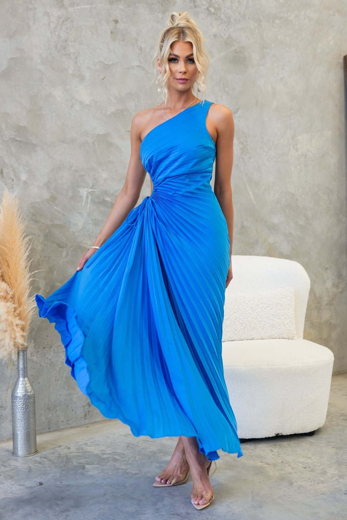 Model in a vibrant blue one-shoulder maxi dress with a cut-out waist, styled with pleats and an elegant flow.