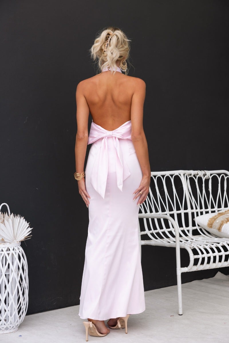 Back view of a woman in a satin pink maxi dress with a bow detail, showcasing a halter neck and open back design.