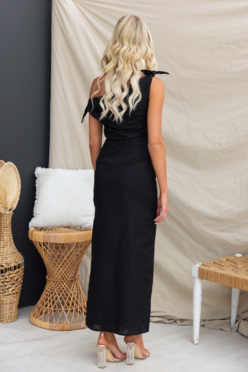 Back view of a woman in a black one-shoulder maxi dress with tie straps, featuring a side split and linen look.